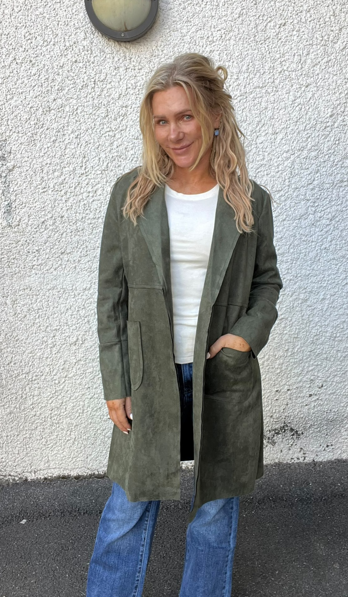 Kvinna med långt blont hår står och ler mot kameran med ena handen i fickan. Hon bär lång olivgrön kappa över vit topp och blå jeans, framför en vit putsad vägg med rund vägglampa ovanför.