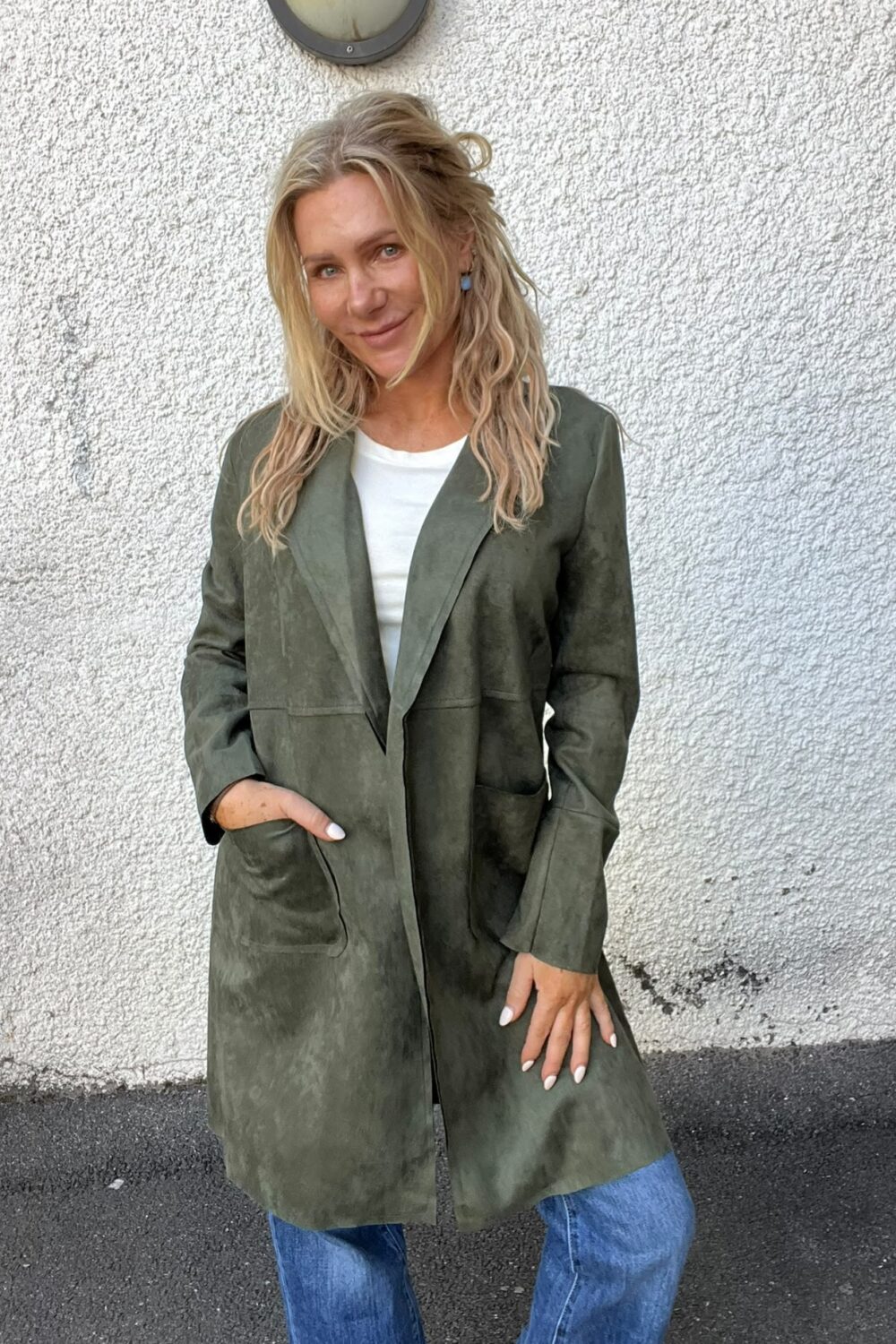 Blond kvinna står och ler mot kameran med ena handen i fickan, klädd i lång olivgrön kappa över vit tröja och blå jeans. Hon poserar framför en vit, grovputsad vägg med rund vägglampa ovanför.