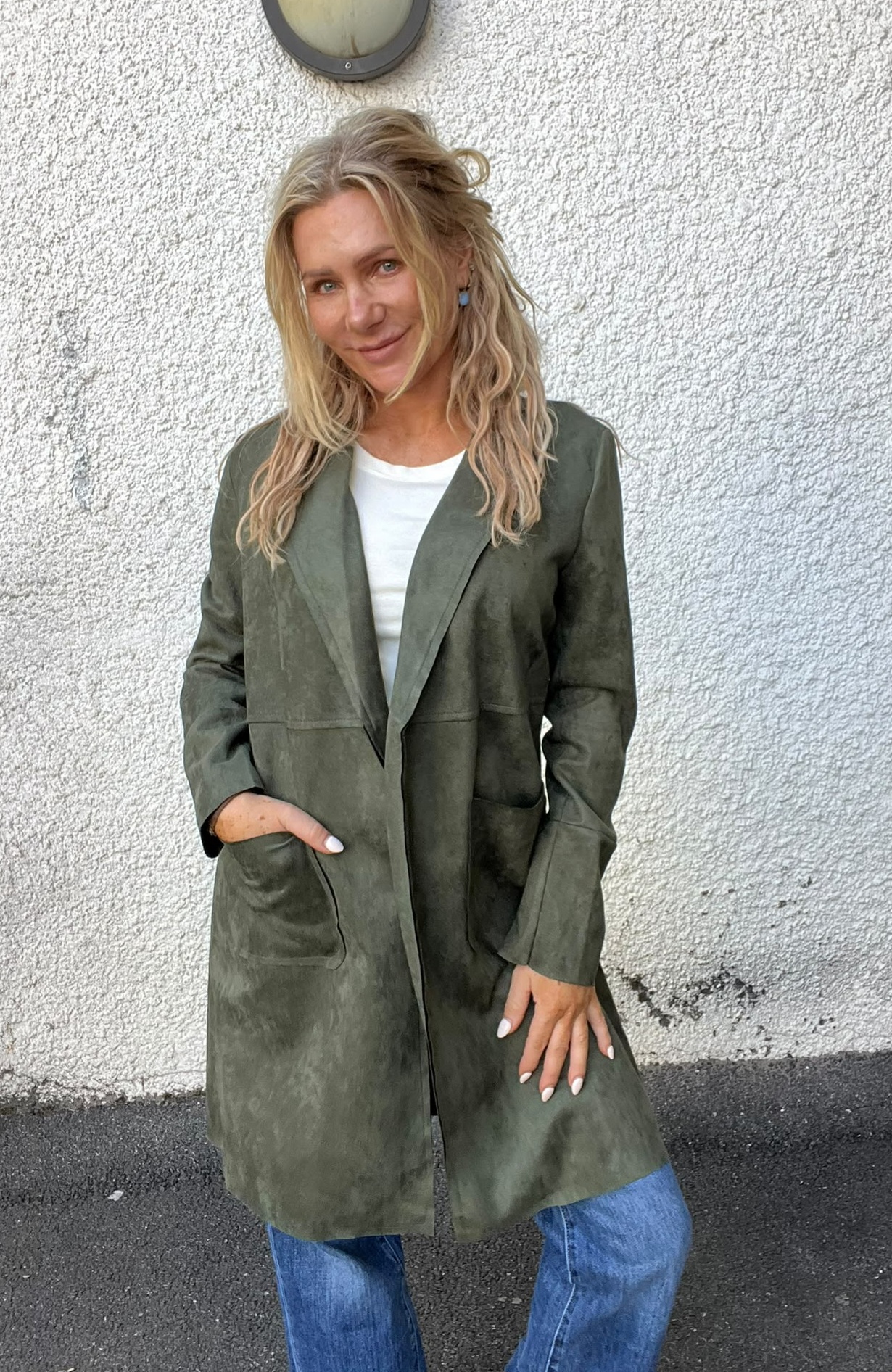 Blond kvinna står och ler mot kameran med ena handen i fickan, klädd i lång olivgrön kappa över vit tröja och blå jeans. Hon poserar framför en vit, grovputsad vägg med rund vägglampa ovanför.