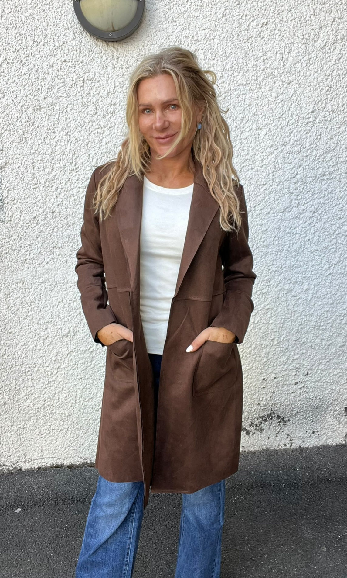 Leende kvinna står avslappnat med händerna i fickorna på en brun, lång kappa, framför en vitputsad vägg utomhus. Hon har långt blont hår, vit topp och blå jeans; en rund vägglampa syns ovanför.