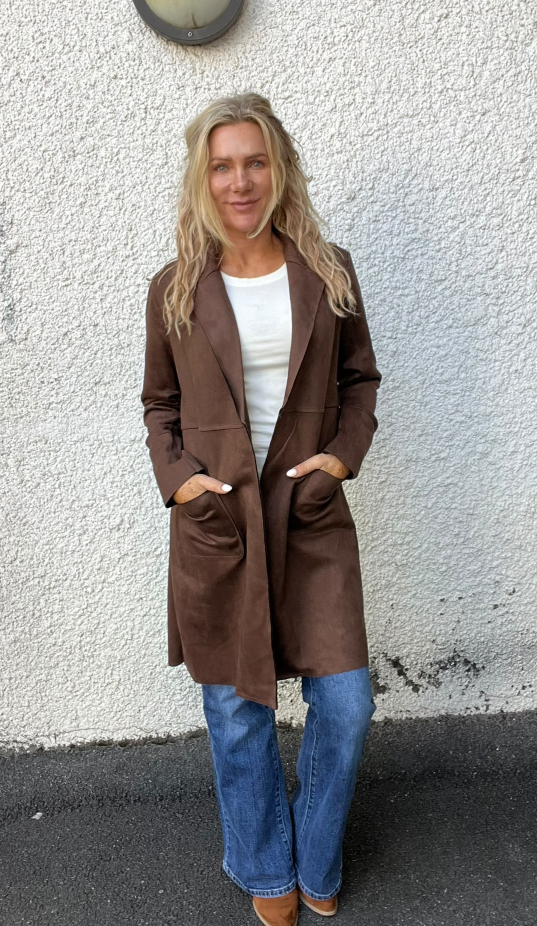 Kvinna med långt blont hår står och ler mot kameran med händerna i fickorna. Hon bär lång brun kappa över vit tröja, blå utsvängda jeans och beige skor, mot en ljus grov vägg utomhus.