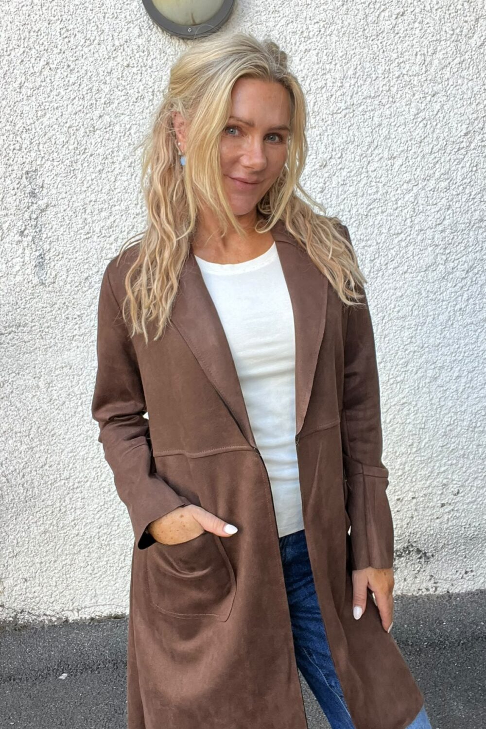 Blond kvinna står och ler lätt mot kameran med ena handen i fickan, klädd i lång brun kappa över vit topp och blå jeans. Hon står utomhus framför en vit, grovputsad vägg med rund vägglampa ovanför.