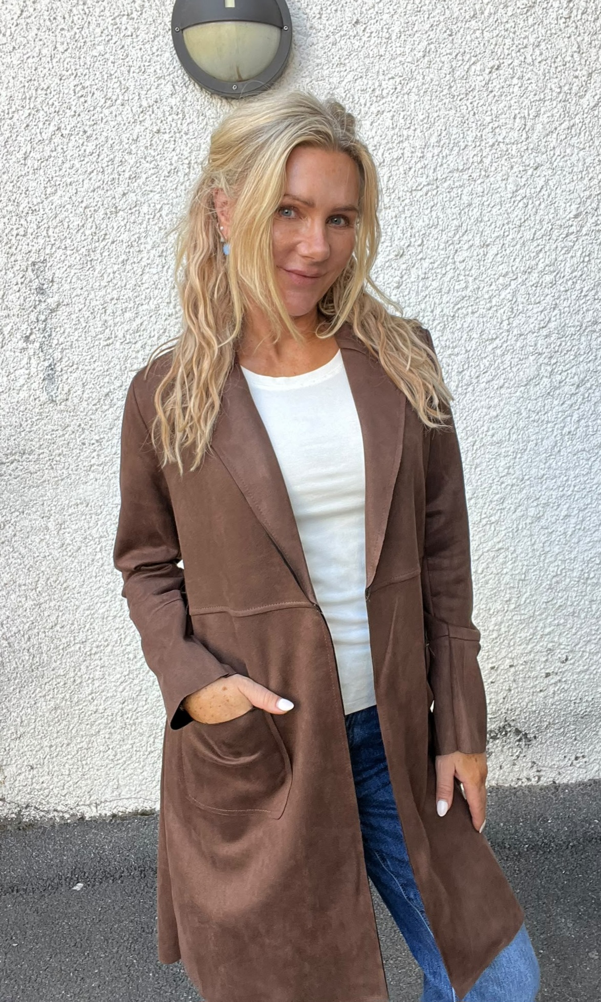 Blond kvinna står och ler lätt mot kameran med ena handen i fickan, klädd i lång brun kappa över vit topp och blå jeans. Hon står utomhus framför en vit, grovputsad vägg med rund vägglampa ovanför.