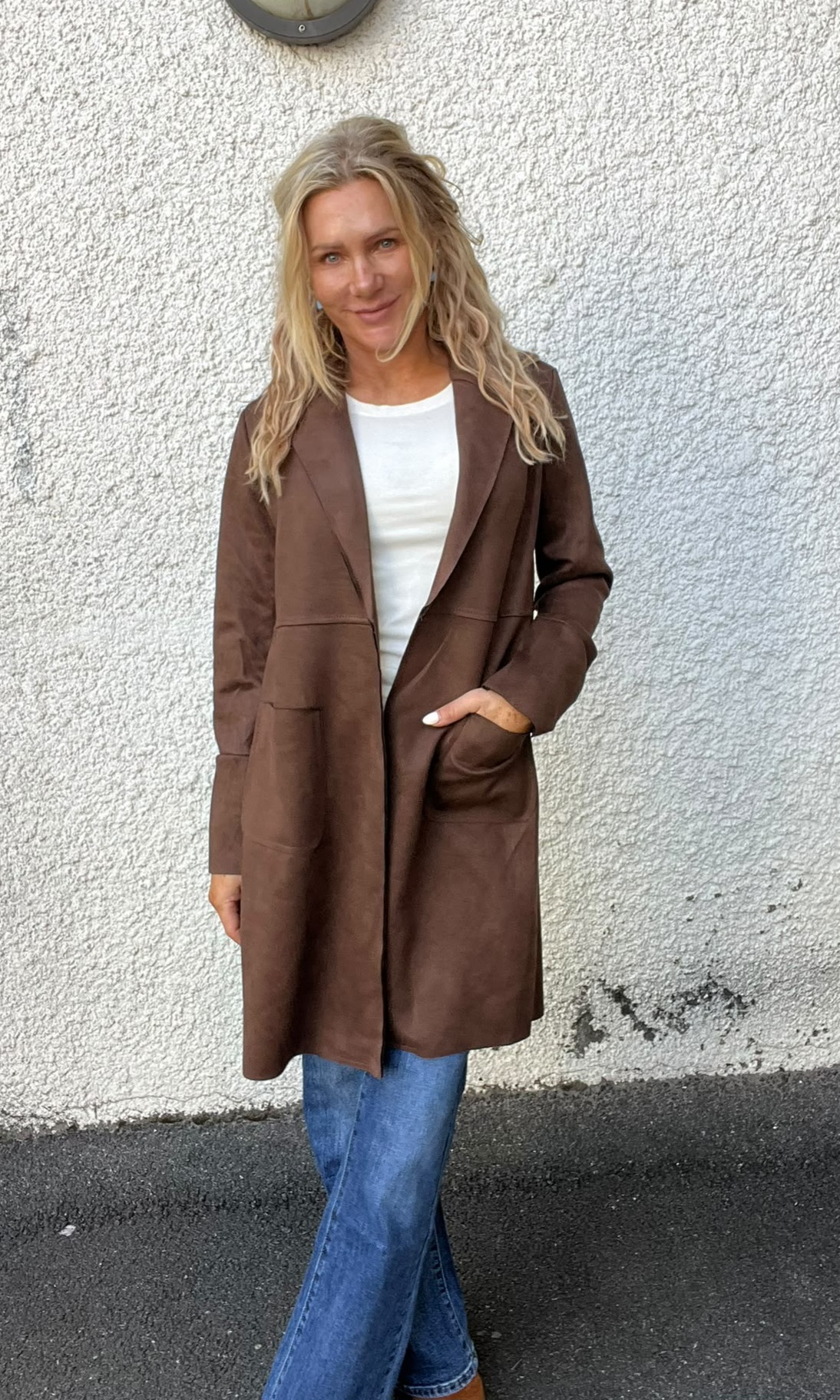 Blond kvinna står och ler mot kameran med ena handen i fickan, klädd i lång brun kappa över vit tröja och blå jeans. Hon står utomhus framför en ljus, grovputsad vägg.