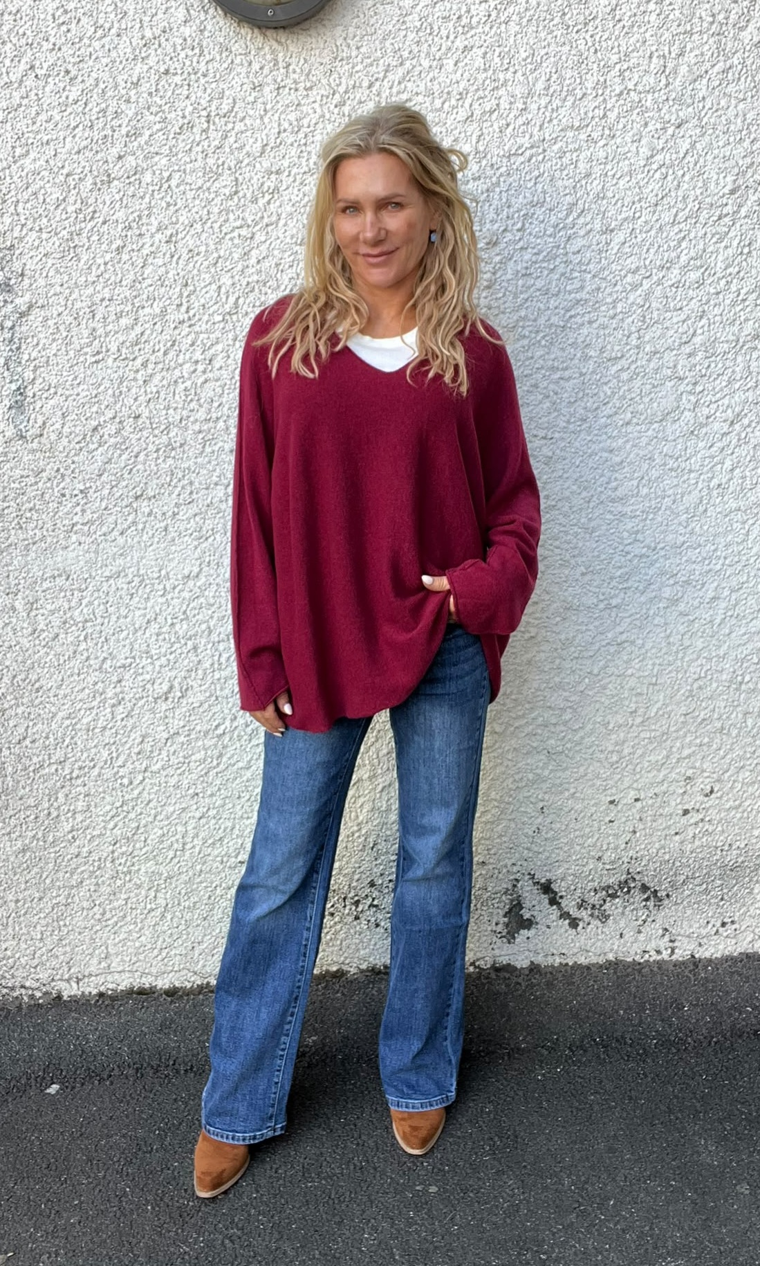 Kvinna med långt blont hår står och ler mot kameran med ena handen i fickan, klädd i vinröd stickad tröja över vit topp, blå jeans och bruna skor, framför en ljus, grovputsad vägg utomhus.