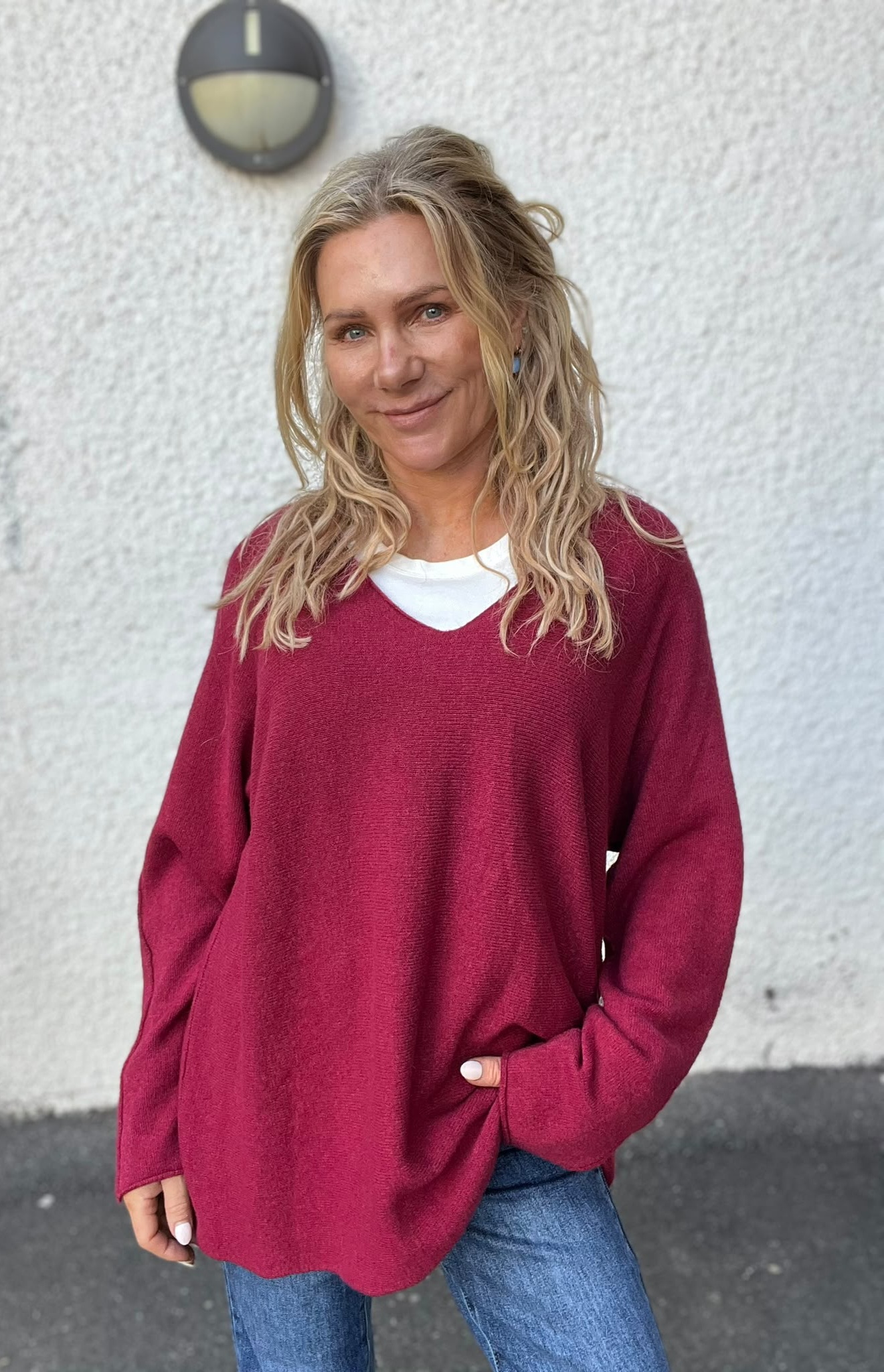 Leende person med långt blont hår står utomhus framför en ljus putsad vägg och tittar mot kameran, klädd i vinröd stickad tröja över vit topp och blå jeans; rund vägglampa syns ovanför.