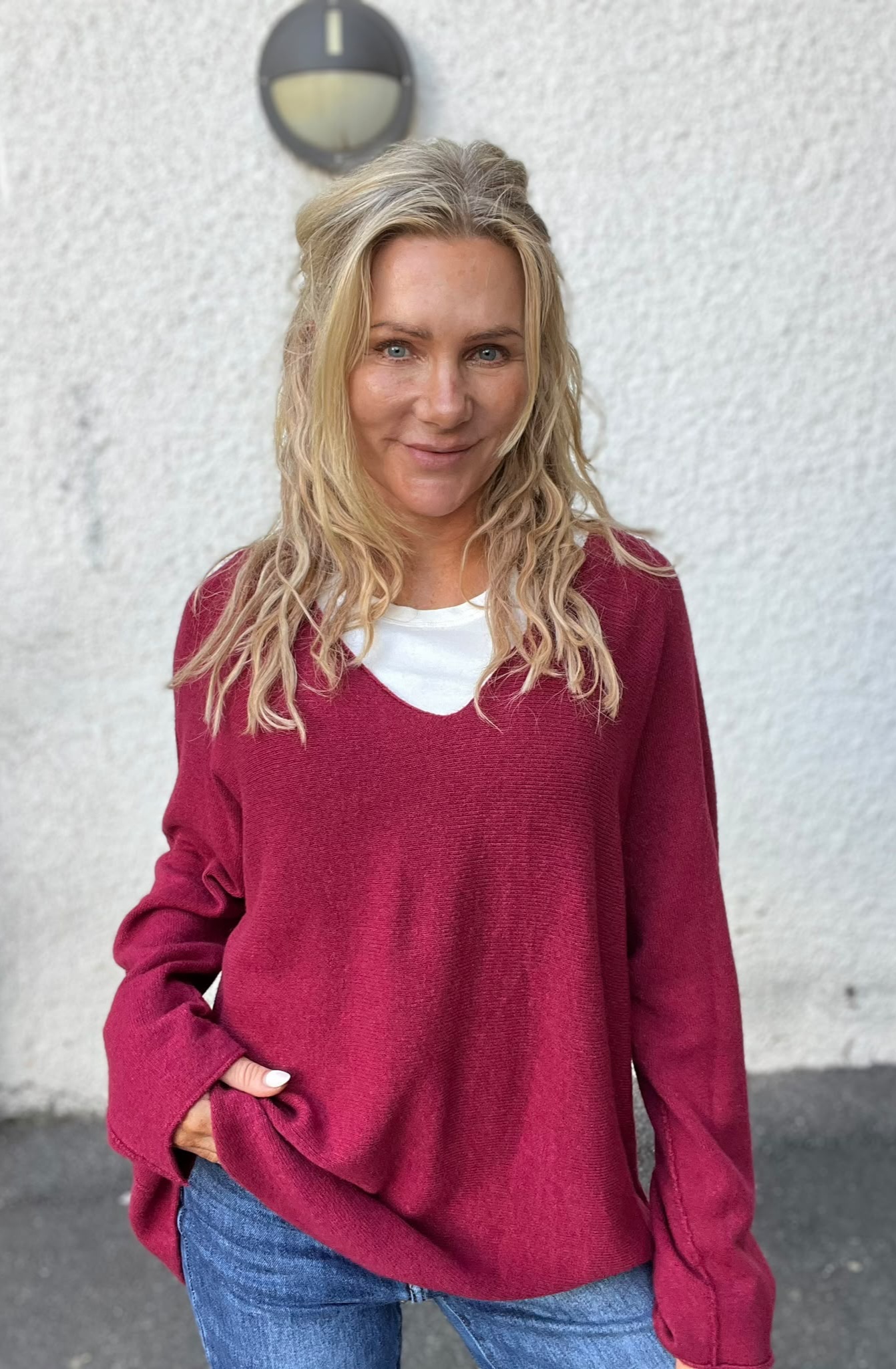 Blond kvinna står och ler mot kameran med ena handen i jeansfickan. Hon bär vinröd stickad tröja över vit topp. Bakom henne syns en ljus putsad vägg med en rund vägglampa ovanför.