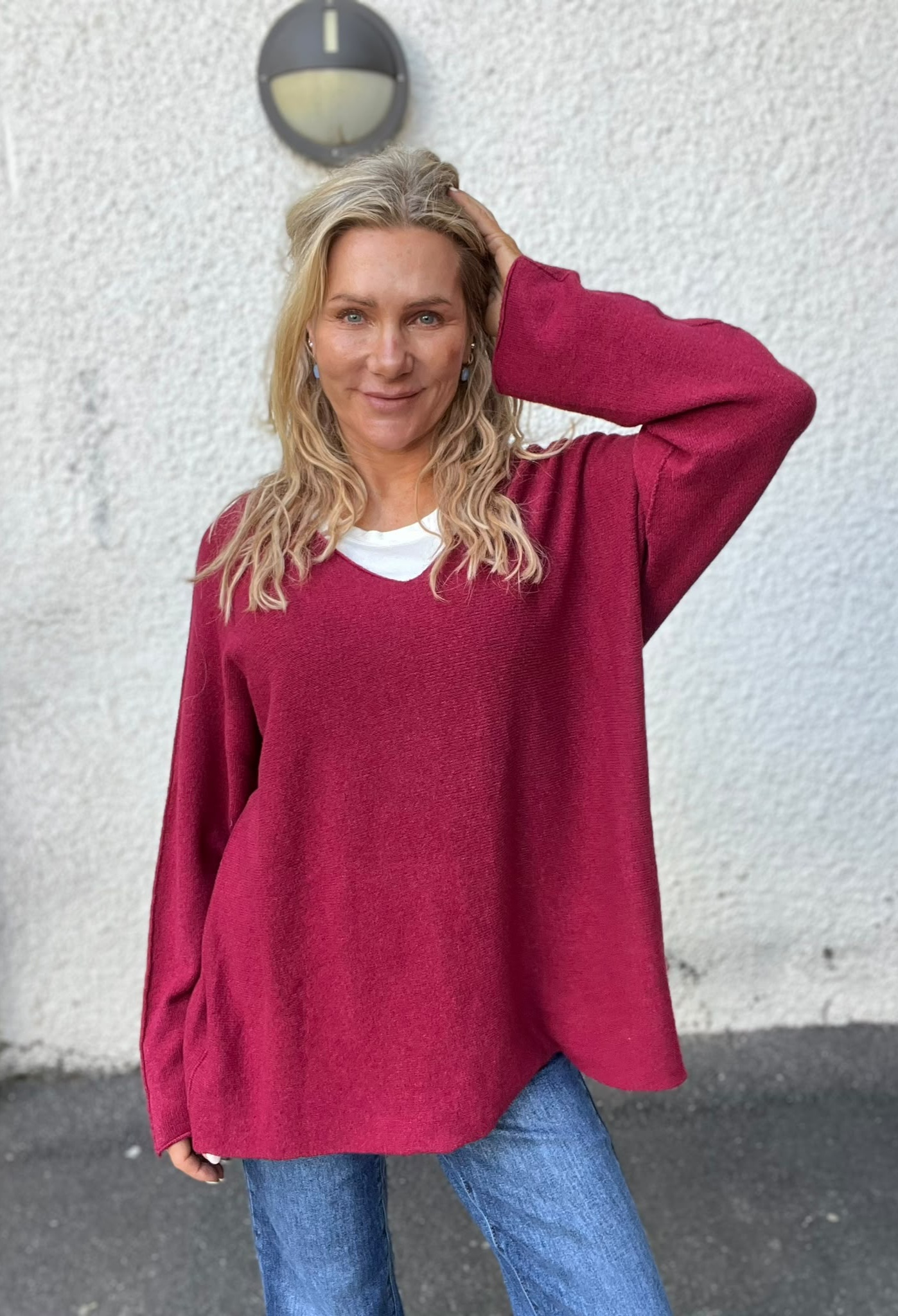 Leende kvinna med långt blont, vågigt hår poserar med ena handen i håret. Hon bär en ceriserosa stickad tröja över vit topp och blå jeans, och står mot en vit putsad vägg med rund vägglampa.