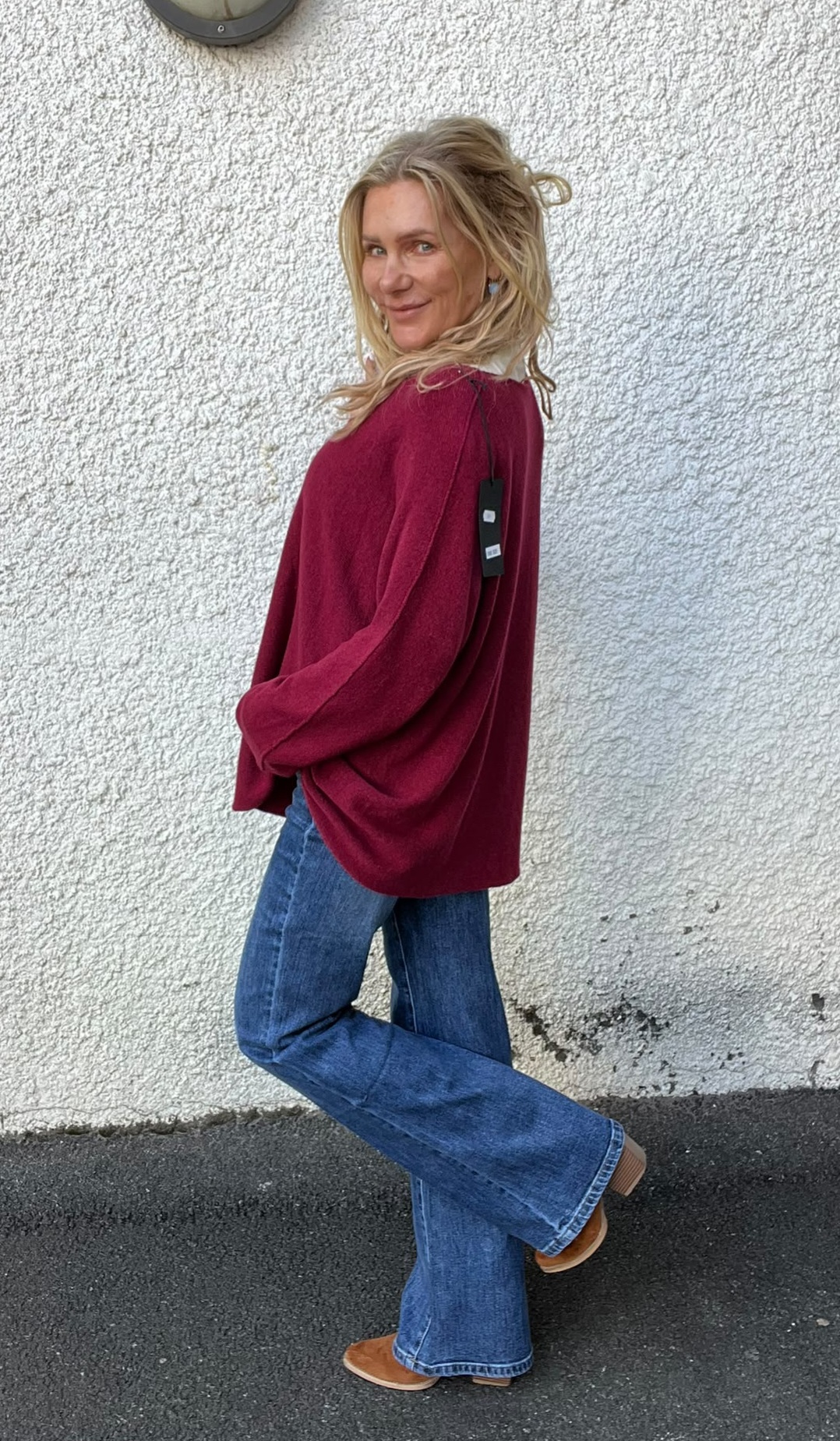 Kvinna står i profil mot en vit, grovputsad vägg och tittar leende mot kameran. Hon visar en vinröd stickad tröja med prislapp bak, blå jeans och bruna skor, ena benet lätt böjt.