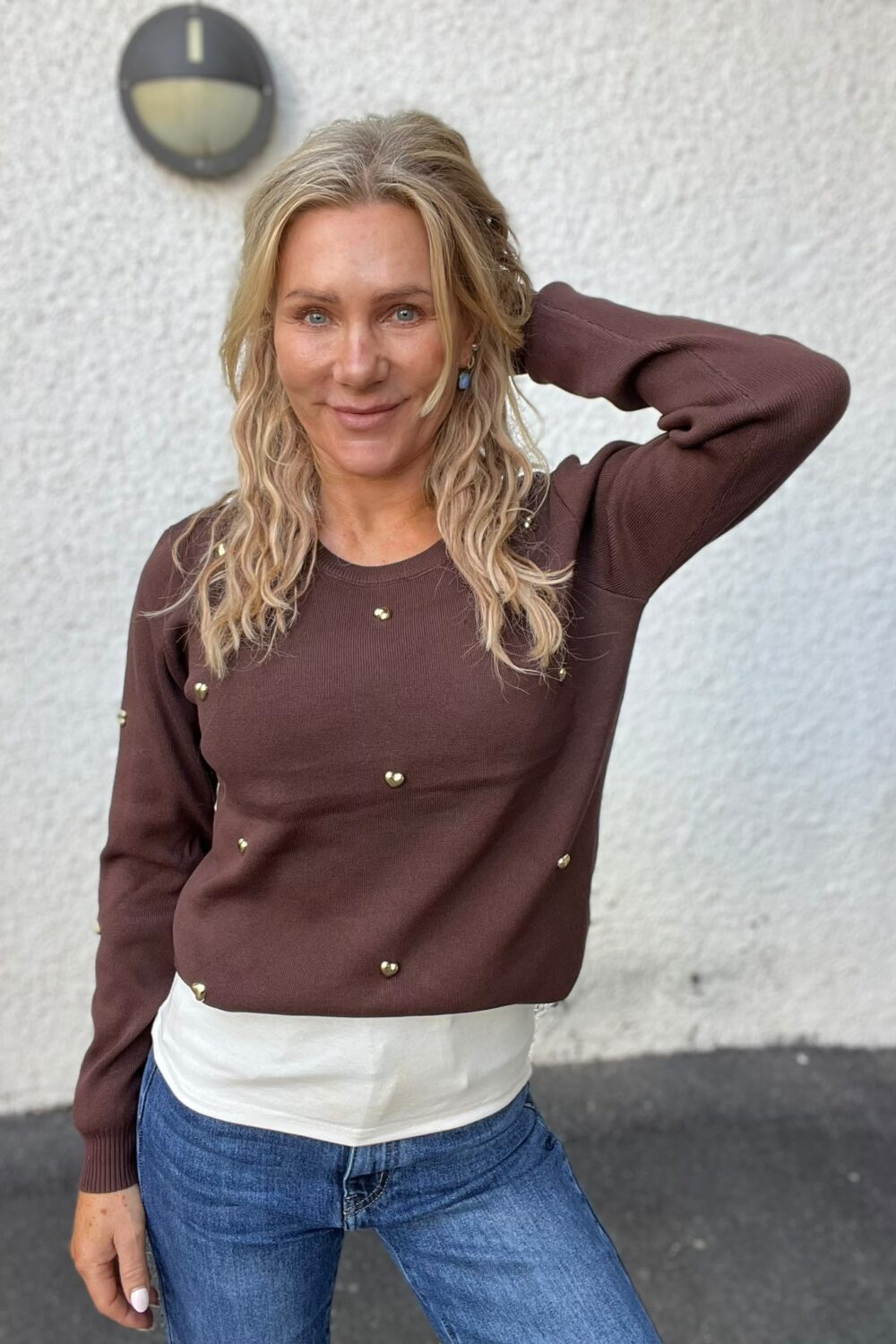 Kvinna med långt blont hår står framför en ljus, grov vägg och ler mot kameran med ena handen bakom huvudet. Hon bär brun tröja med guldfärgade knappar och blå jeans; en rund vägglampa syns bakom.