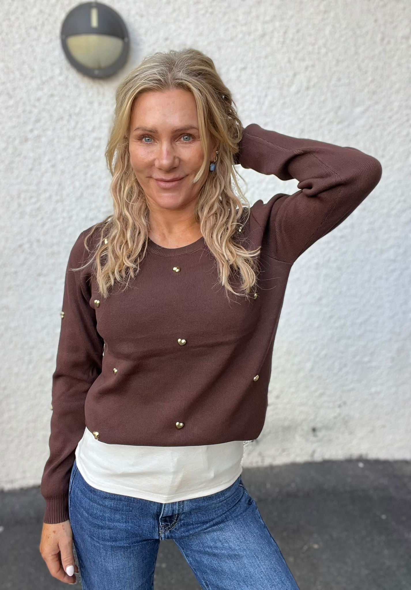 Kvinna med långt blont hår står framför en ljus, grov vägg och ler mot kameran med ena handen bakom huvudet. Hon bär brun tröja med guldfärgade knappar och blå jeans; en rund vägglampa syns bakom.