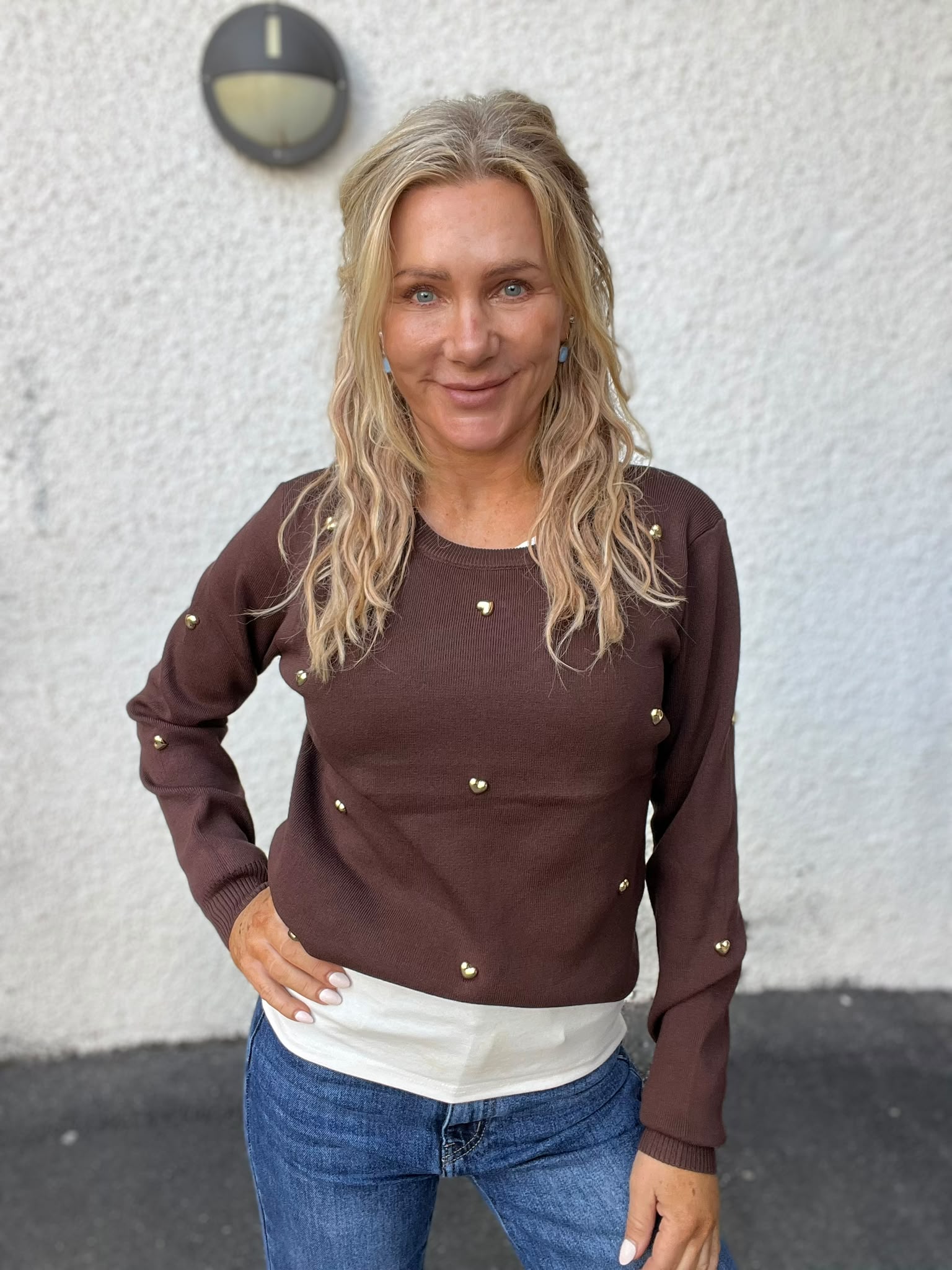 Leende kvinna med långt blont hår står vänd mot kameran med ena handen på höften, klädd i brun tröja med guldfärgade knappar och jeans. Bakom henne syns en ljus vägg med en rund vägglampa.