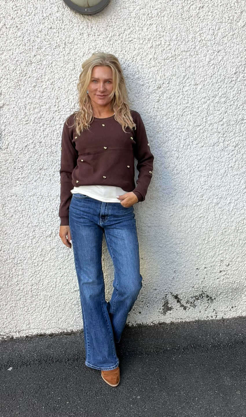 Kvinna med långt blont hår står lutad mot en vit, grovputsad vägg och tittar mot kameran. Hon har brun tröja med små dekorationer, blå jeans och bruna boots; ena handen i fickan, benen korsade.