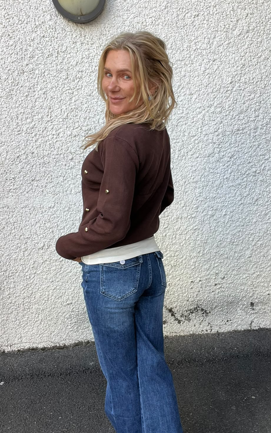Blond kvinna står i profil och vänder sig leende mot kameran, med händerna i fickorna. Hon bär brun tröja med knappar på ärmen och blå jeans, framför en vit putsad vägg med rund vägglampa.