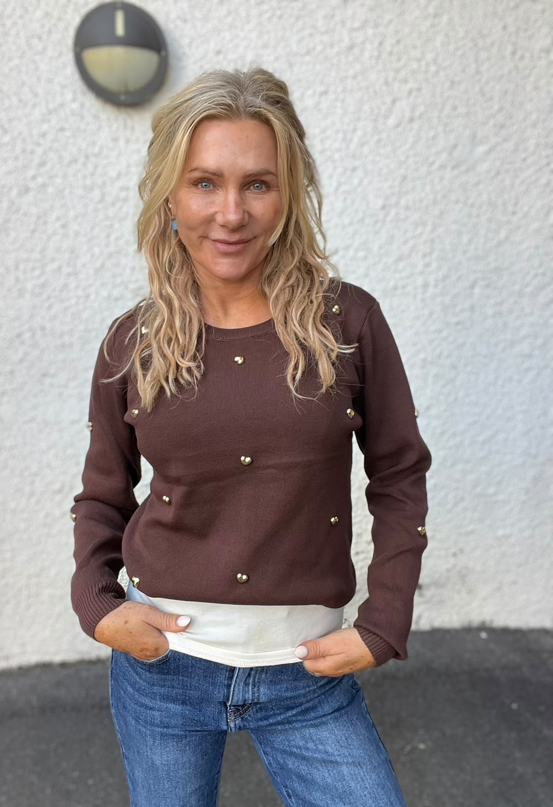 Blond kvinna står och ler mot kameran med händerna i jeansfickorna. Hon bär brun tröja med små guldfärgade nitar och vit topp under, framför en ljus putsad vägg med rund vägglampa.