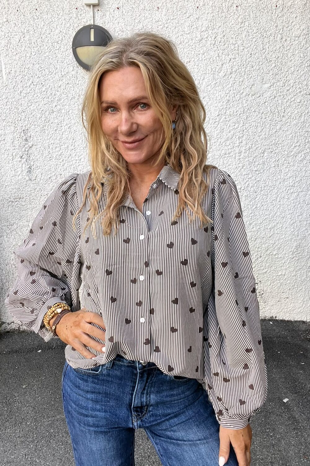 Blond kvinna står och ler mot kameran med ena handen på höften. Hon bär en randig skjorta med små hjärtmönster och blå jeans, framför en vitputsad vägg med rund vägglampa.