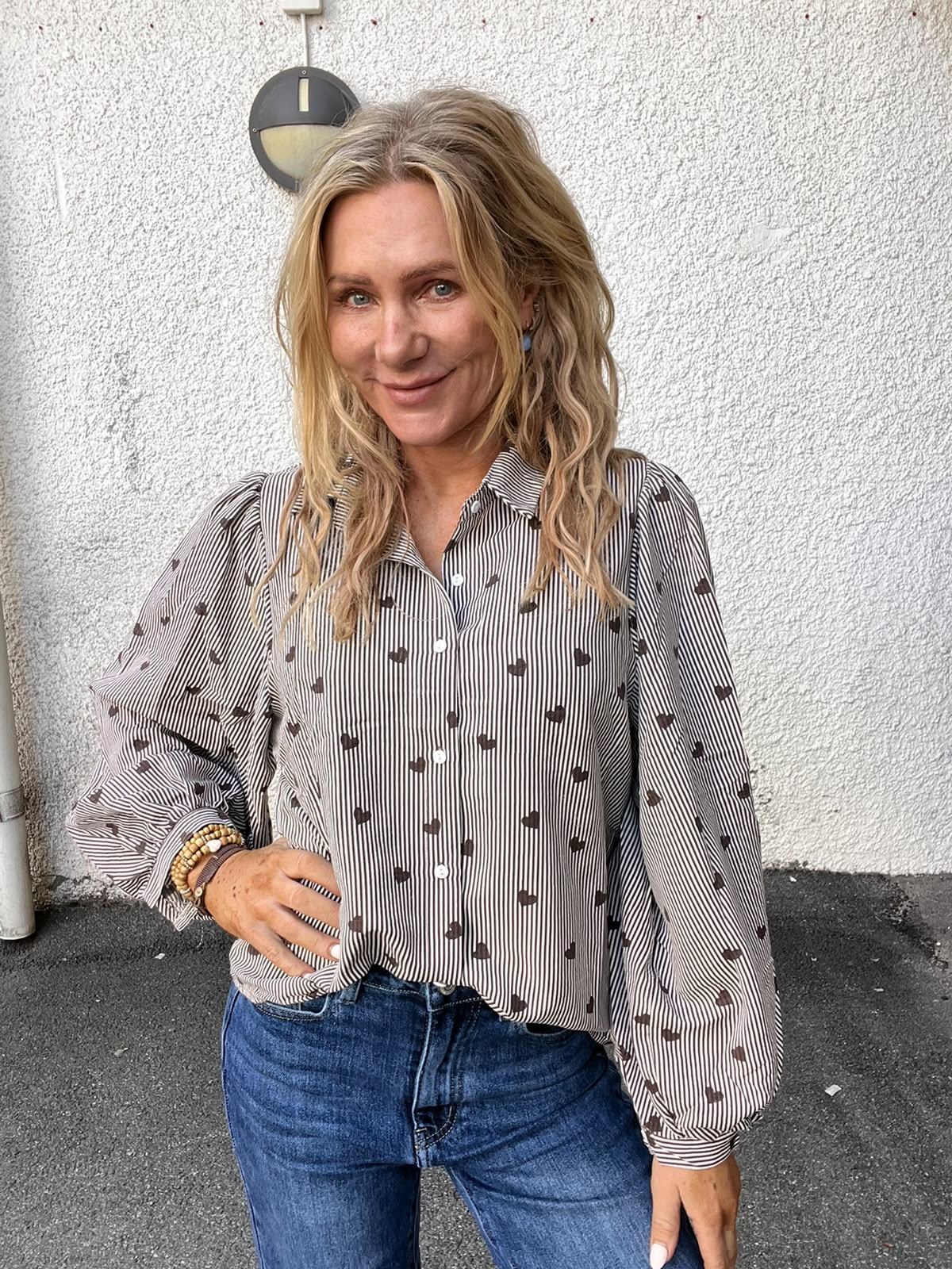 Blond kvinna står och ler mot kameran med ena handen på höften. Hon bär en randig skjorta med små hjärtmönster och blå jeans, framför en vitputsad vägg med rund vägglampa.