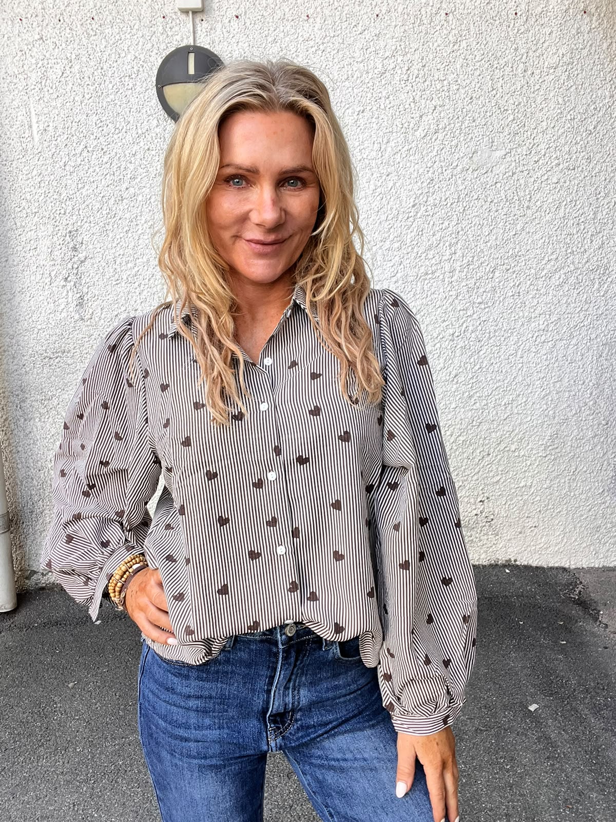 Blond kvinna står och ler mot kameran med ena handen på höften. Hon bär randig skjorta med små hjärtan, blå jeans och armband. Bakgrunden är en ljus, grov vägg med en rund vägglampa.