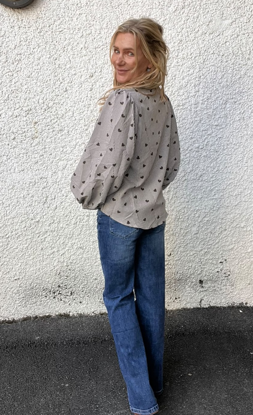 Kvinna står utomhus mot en vit, grovputsad vägg och vänder sig om med ett leende. Hon har blondt hår, bär en gråblus med små mörka motiv och vida ärmar samt blå jeans.