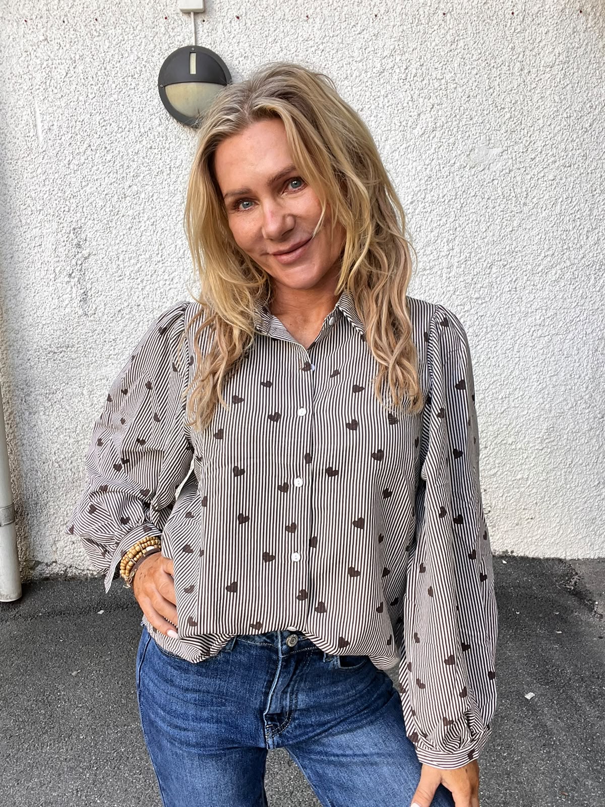 Kvinna med långt blont hår ler och poserar med handen på höften framför en vit putsad vägg. Hon bär blå jeans och en randig skjorta med små hjärtan samt flera armband.