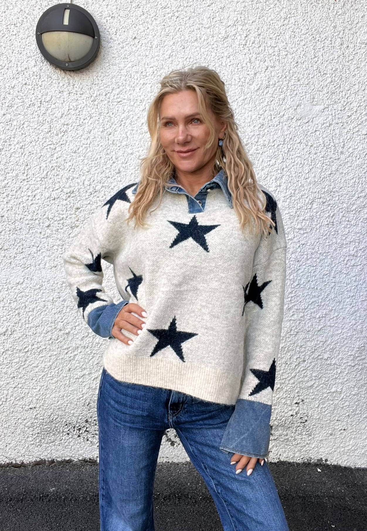 Blond kvinna står med ena handen på höften och ler lätt mot kameran. Hon bär jeans, jeansskjorta och en ljus stickad tröja med mörka stjärnor. Bakgrund: vit putsad vägg med rund vägglampa.