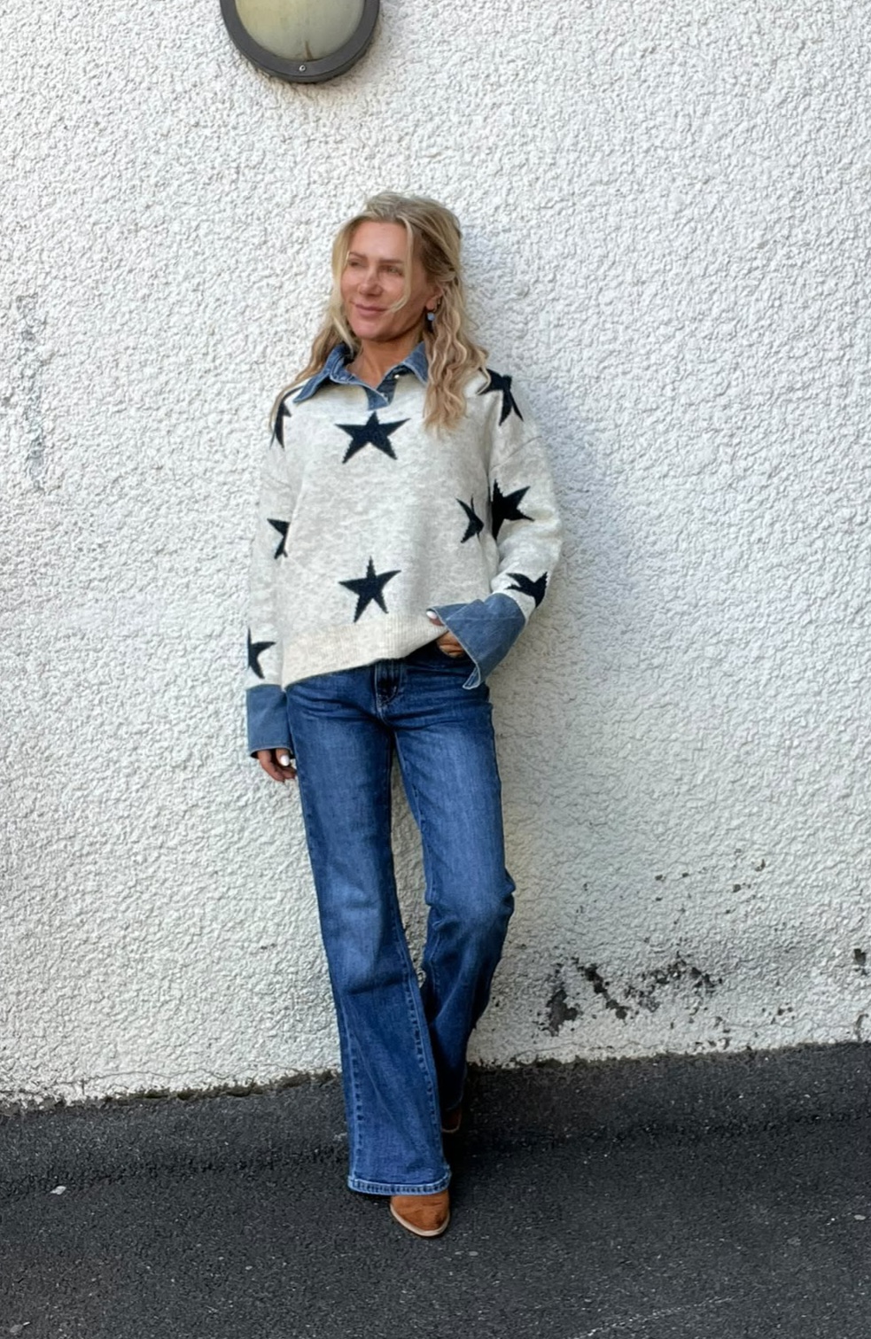 Blond kvinna står avslappnat lutad mot en vit, grovputsad vägg och ser åt sidan. Hon har en ljus tröja med stora svarta stjärnor över en jeansskjorta, blå jeans och bruna skor.