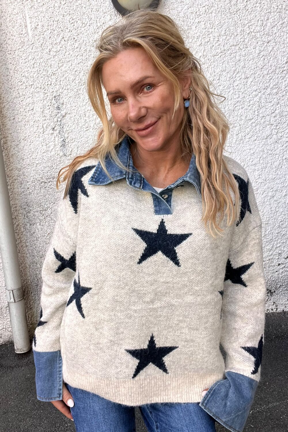 Blond kvinna ler mot kameran och lutar sig lätt mot en vit, grovputsad vägg utomhus. Hon bär en ljus tröja med stora mörka stjärnor över en jeansskjorta och blå örhängen.