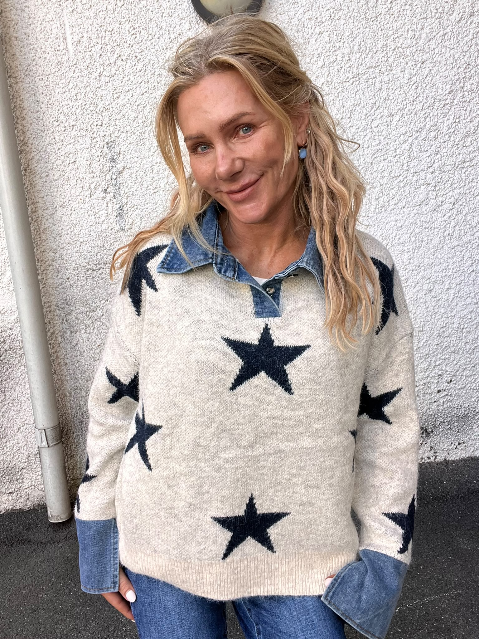 Blond kvinna ler mot kameran och lutar sig lätt mot en vit, grovputsad vägg utomhus. Hon bär en ljus tröja med stora mörka stjärnor över en jeansskjorta och blå örhängen.