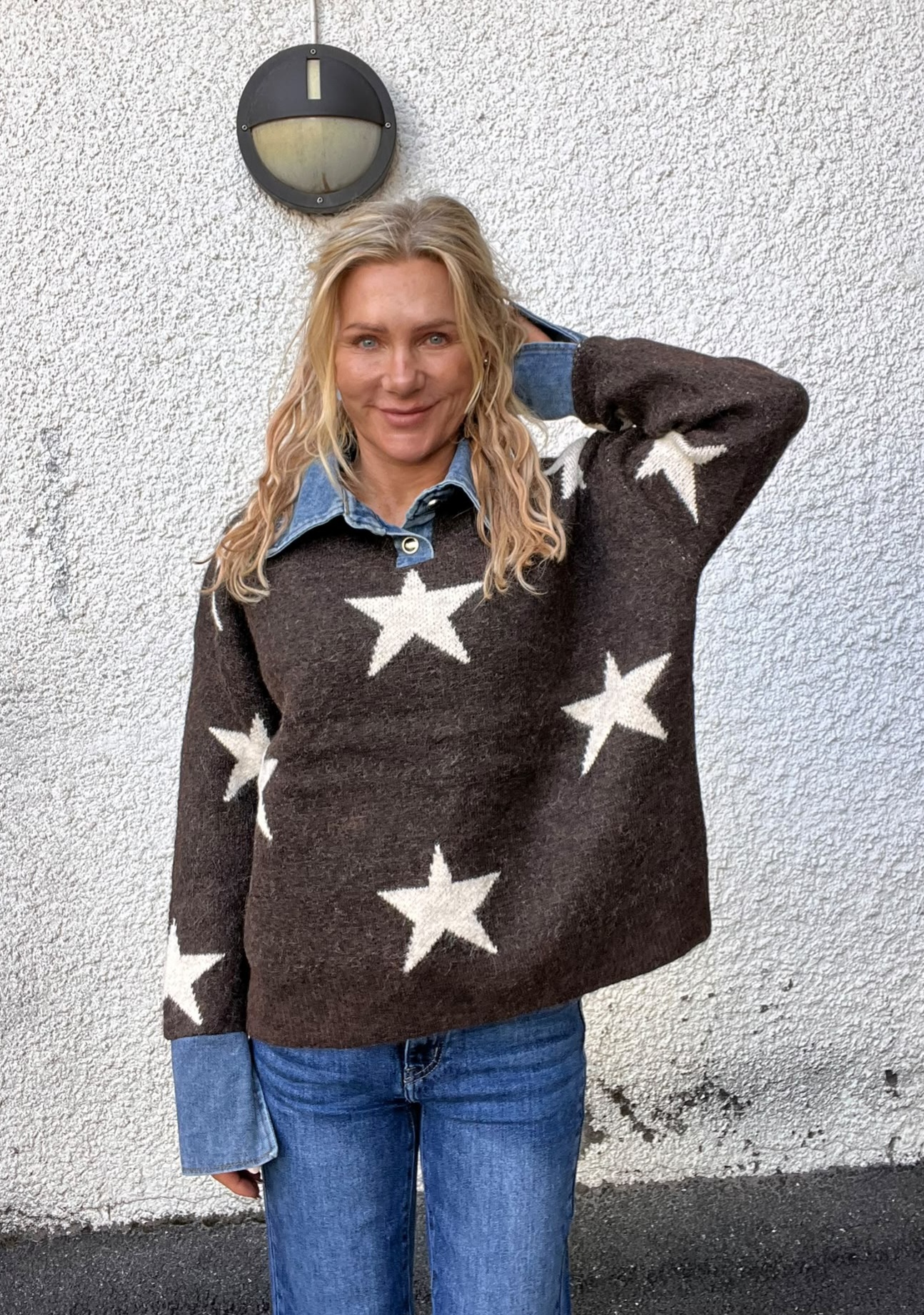 Blond kvinna står och ler mot kameran med ena handen bakom huvudet. Hon bär brun stickad tröja med stora vita stjärnor över en jeansskjorta och blå jeans, framför en vit putsad vägg med rund vägglampa.