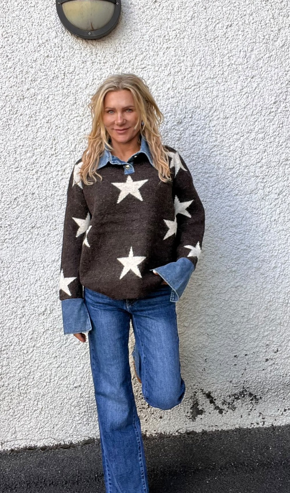 Kvinna med långt blont hår står lutad mot en vit putsad vägg och ler mot kameran. Hon har en mörk tröja med stora vita stjärnor, denimskjorta under och blå jeans, en hand i fickan.