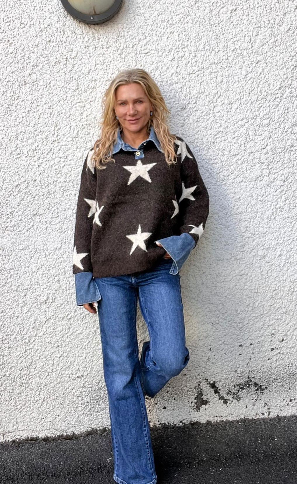 Blond kvinna står lutad mot en vit, grovputsad vägg och ler mot kameran, med ett ben böjt. Hon bär mörk stickad tröja med stora vita stjärnor över jeansskjorta samt blå jeans.