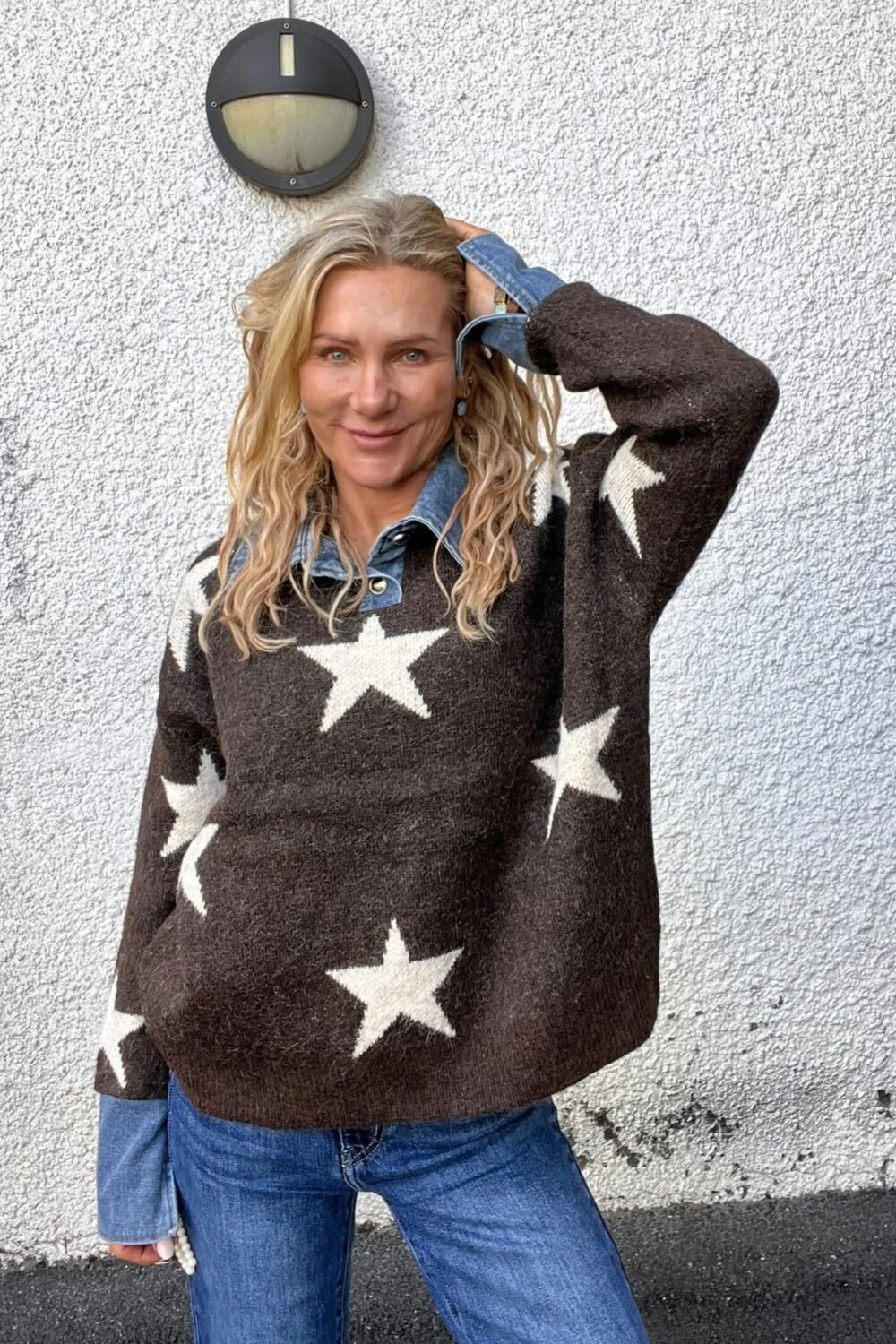 Kvinna med långt blont hår står mot en vit, grovputsad vägg och ler mot kameran, med ena handen i håret. Hon bär brun stickad tröja med vita stjärnor över jeansskjorta och blå jeans; vägglampa ovanför.