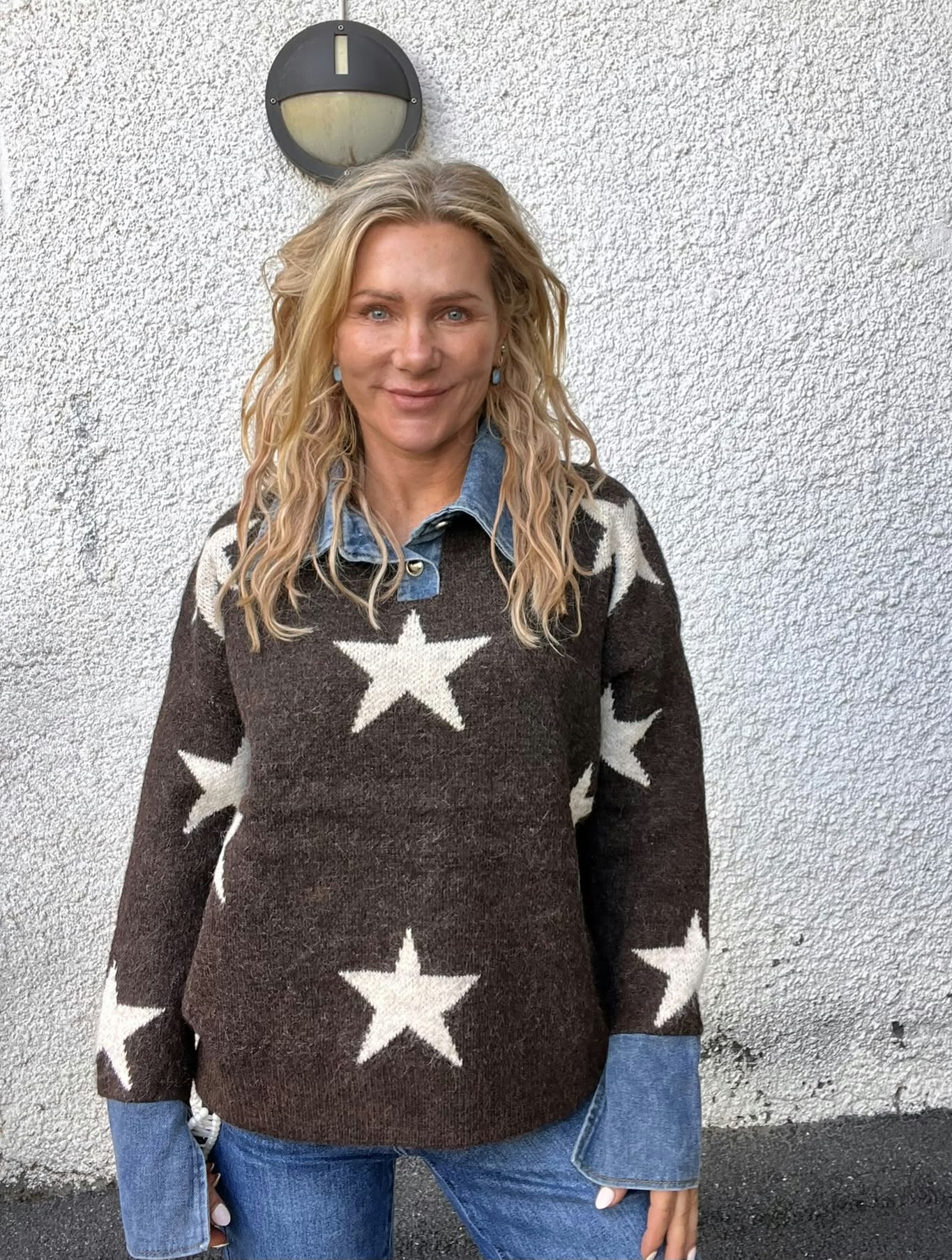 Kvinna med långt blont hår står och ler mot kameran framför en ljus, grovputsad vägg. Hon bär brun stickad tröja med stora vita stjärnor över en blå jeansskjorta och jeans; vägglampa ovanför.