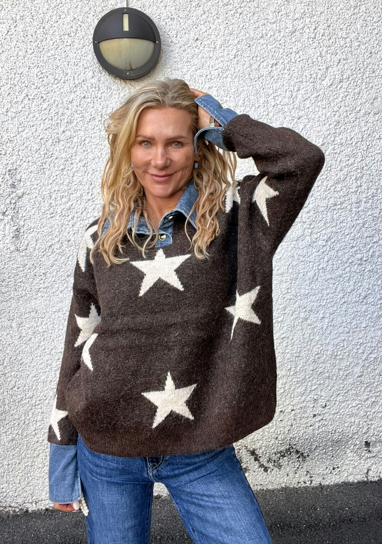 Kvinna med långt blont hår står mot en vit, grovputsad vägg och ler mot kameran, med ena handen i håret. Hon bär brun stickad tröja med vita stjärnor över jeansskjorta och blå jeans; vägglampa ovanför.
