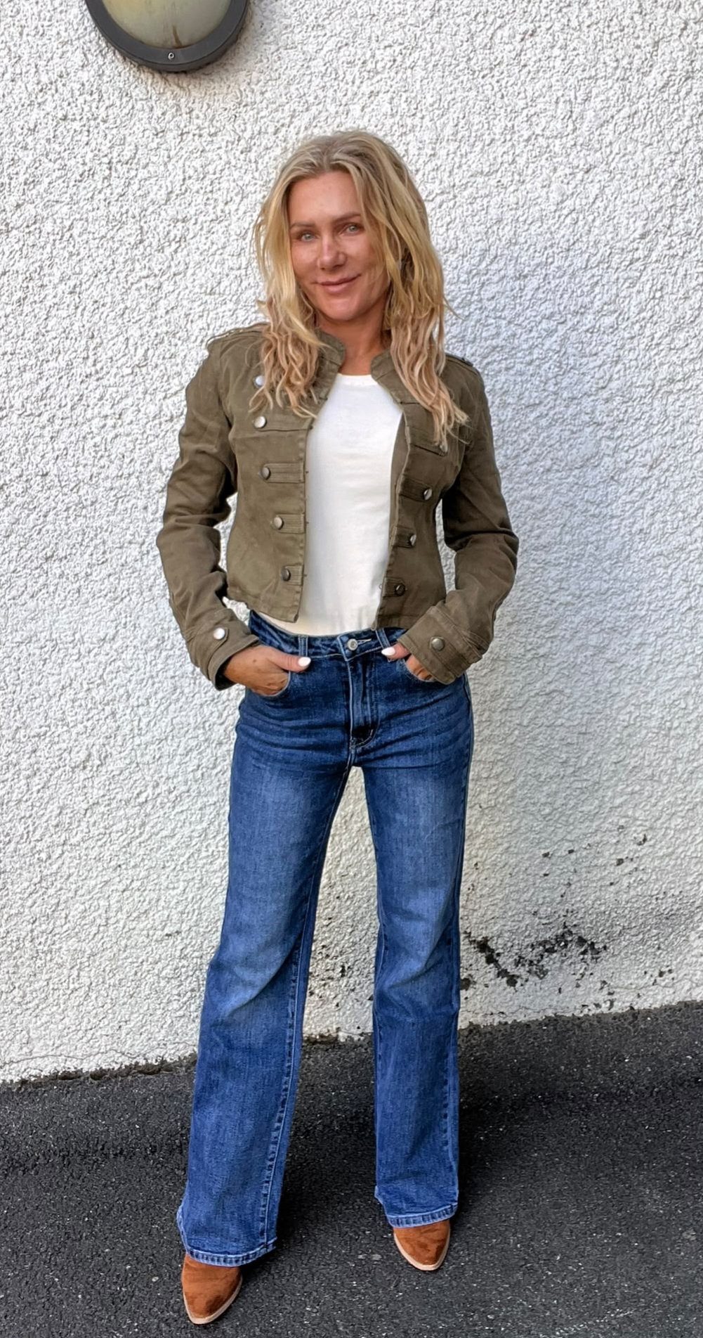 Blond kvinna står med händerna i jeansfickorna och ler mot kameran, framför en vit, grovputsad vägg utomhus. Hon bär olivgrön jacka med knappar, vit topp, blå utsvängda jeans och bruna skor.