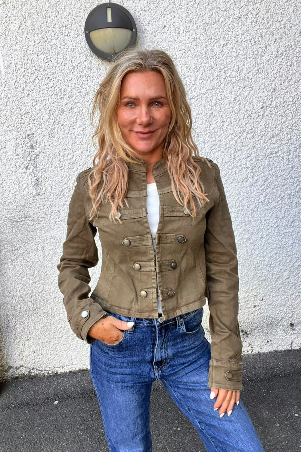 Leende kvinna med långt blont hår står och poserar med ena handen i jeansfickan, klädd i olivgrön jacka och blå jeans. Hon står framför en vit putsad vägg med en rund vägglampa ovanför.