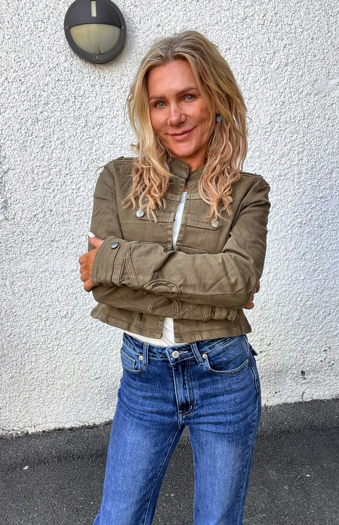 Leende kvinna med långt blondt hår står med armarna i kors och tittar mot kameran. Hon bär olivgrön jacka och blå jeans, framför en vit putsad vägg med rund utomhuslampa ovanför.