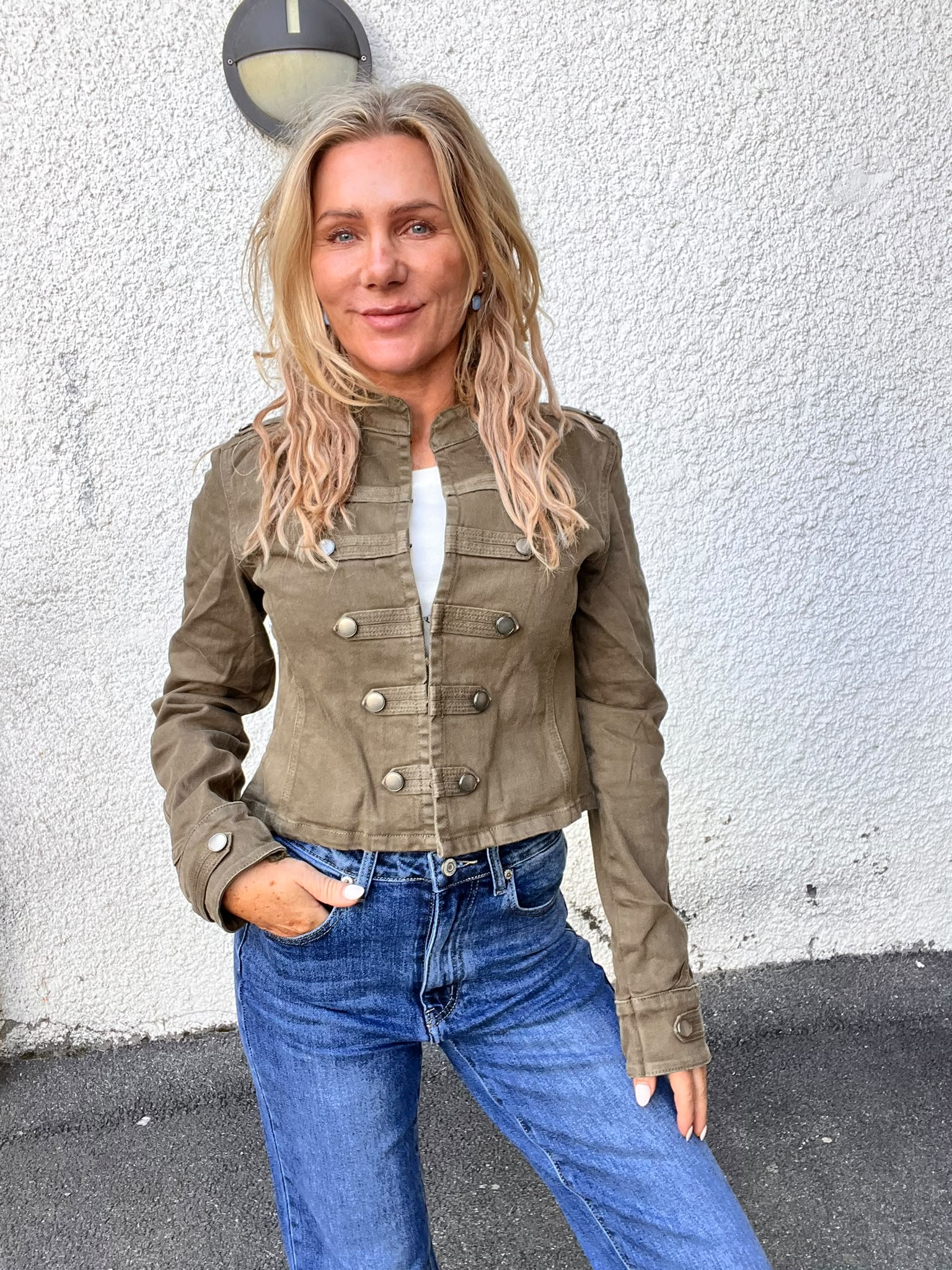 Kvinna med långt blont hår står och ler mot kameran med ena handen i jeansfickan. Hon bär en olivgrön jacka med stora knappar och står framför en ljus, grovputsad vägg med rund vägglampa.