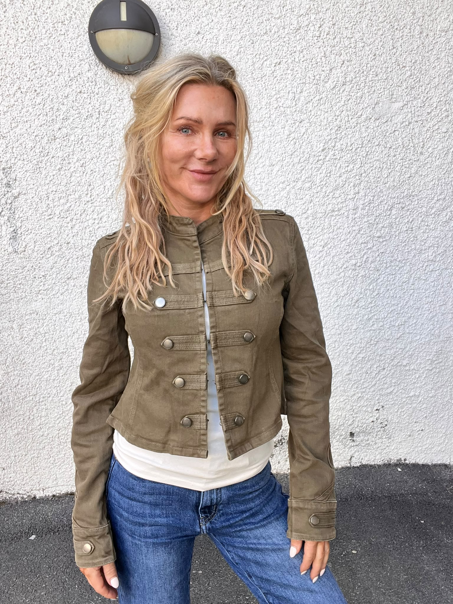 Kvinna med långt blont hår står och ler mot kameran, klädd i olivgrön jacka med knappar, vit tröja och blå jeans. Hon lutar lätt mot en vit, grovputsad vägg med en rund vägglampa ovanför.