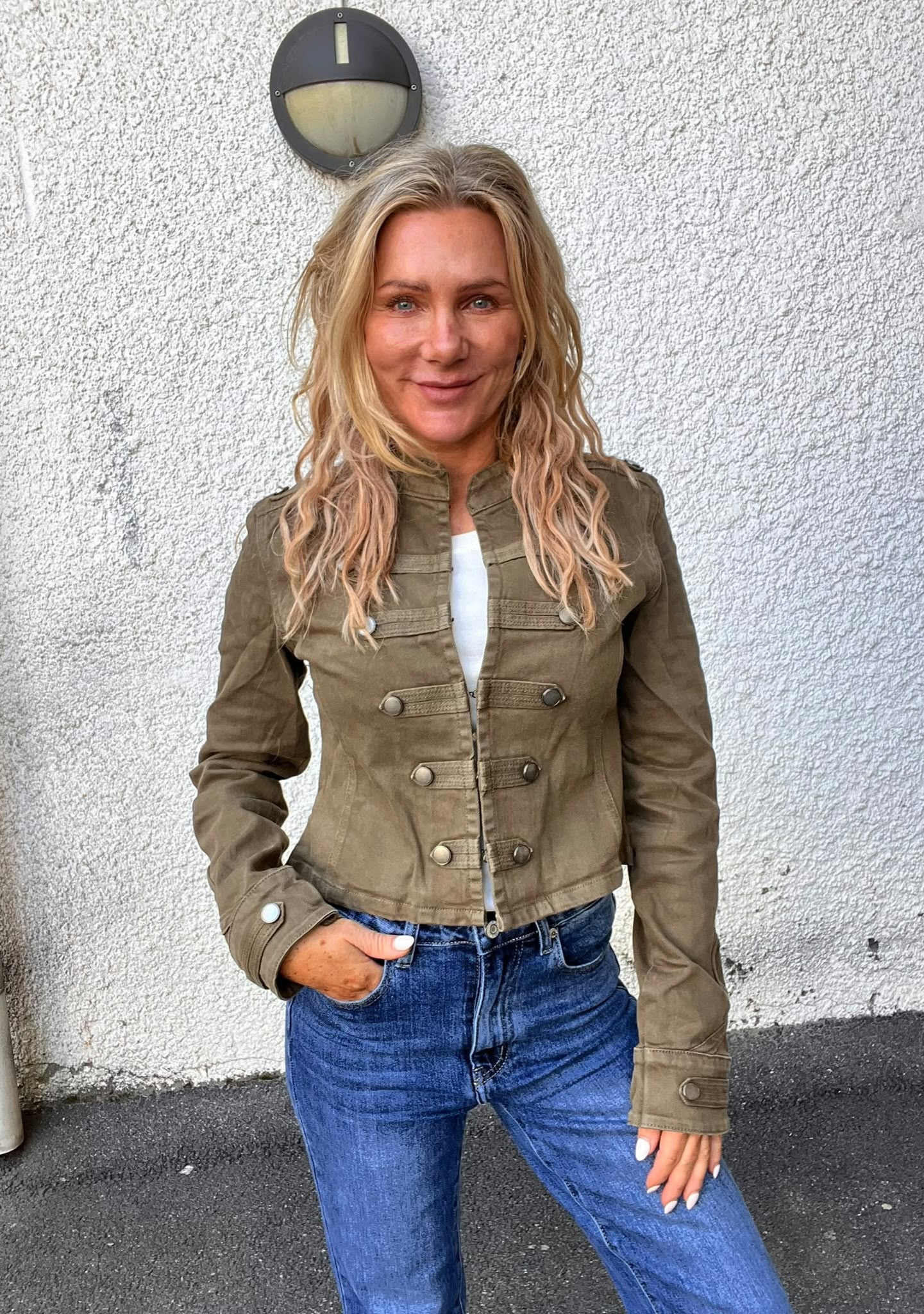 Leende kvinna med långt blont hår står och poserar med ena handen i jeansfickan, klädd i olivgrön jacka och blå jeans. Hon står framför en vit putsad vägg med en rund vägglampa ovanför.
