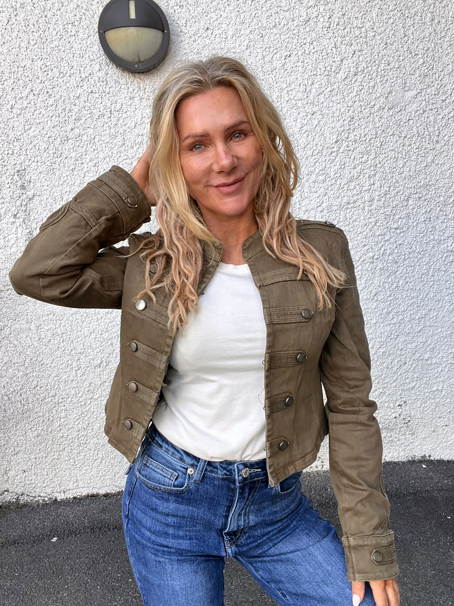 Leende kvinna med långt blont hår poserar med ena handen bakom huvudet. Hon bär olivgrön jacka över vit topp och blå jeans, och står framför en vit grovputsad vägg med rund vägglampa.