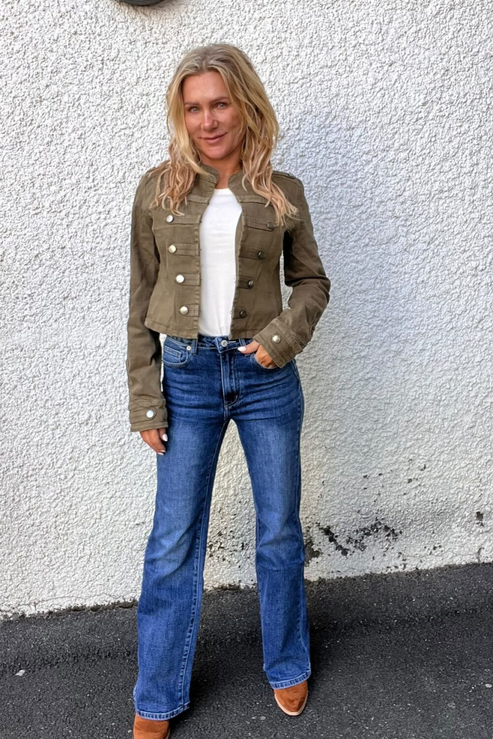Blond kvinna står och poserar avslappnat med ena handen i jeansfickan, framför en vit, grovputsad vägg. Hon bär olivgrön jacka över vit topp, blå utsvängda jeans och bruna skor.