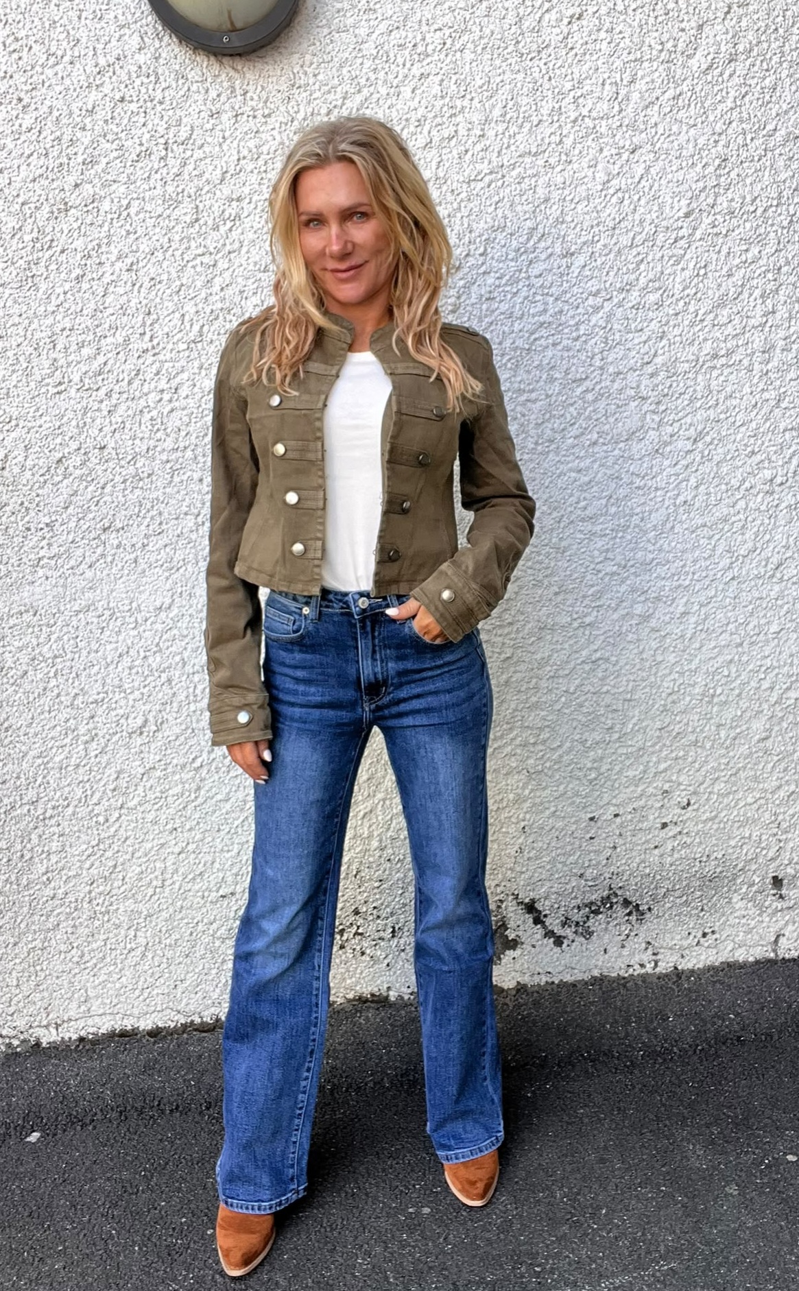 Kvinna med långt blont hår står avslappnat med ena handen i fickan och ler mot kameran, klädd i olivgrön kort jacka, vit topp, blå jeans och bruna stövlar, framför en vitputsad vägg utomhus.