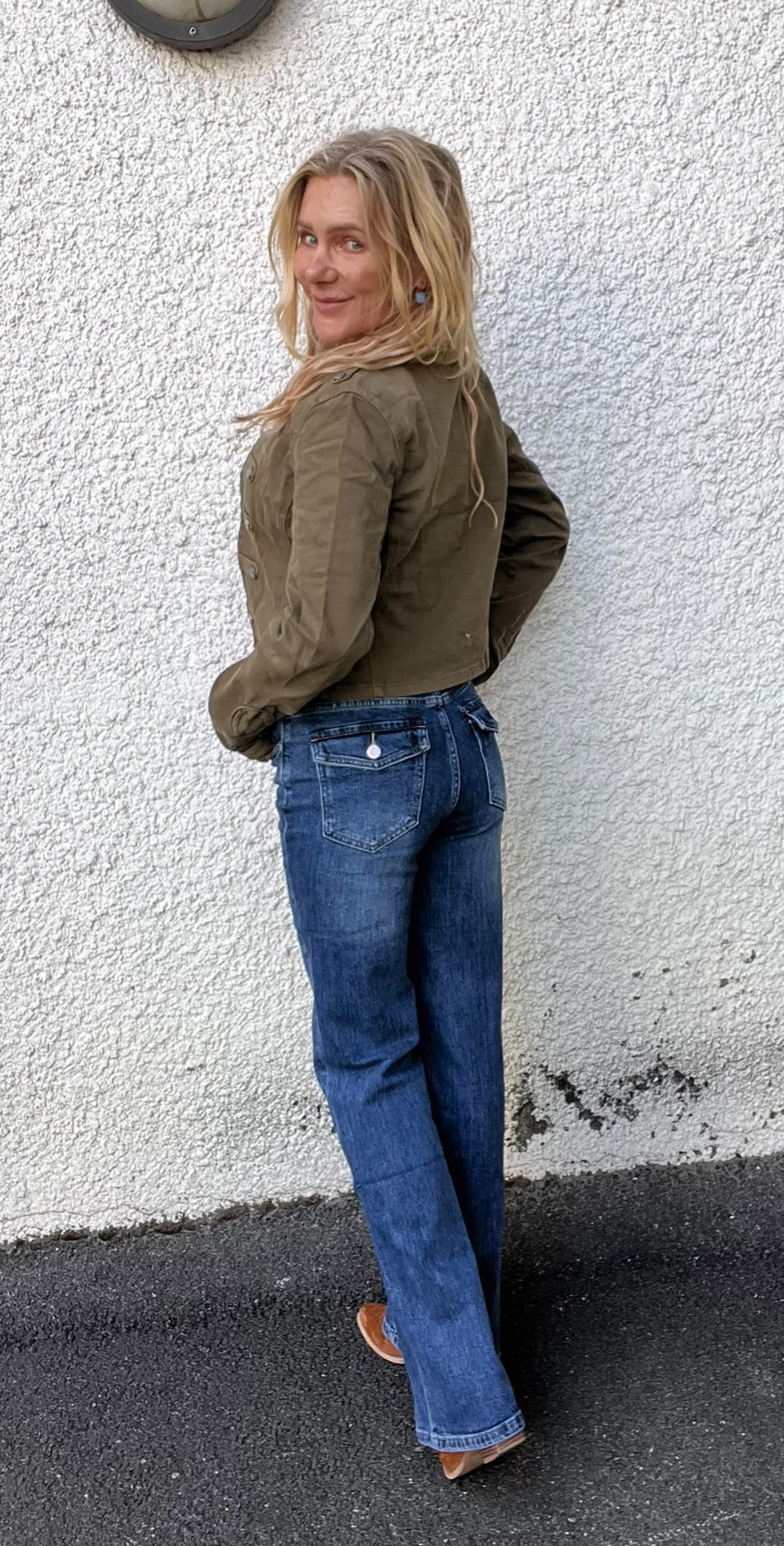 Kvinna med långt blont hår står vänd i profil och tittar leende över axeln mot kameran. Hon bär olivgrön jacka, blå utsvängda jeans och bruna skor, framför en vit grovputsad vägg utomhus.