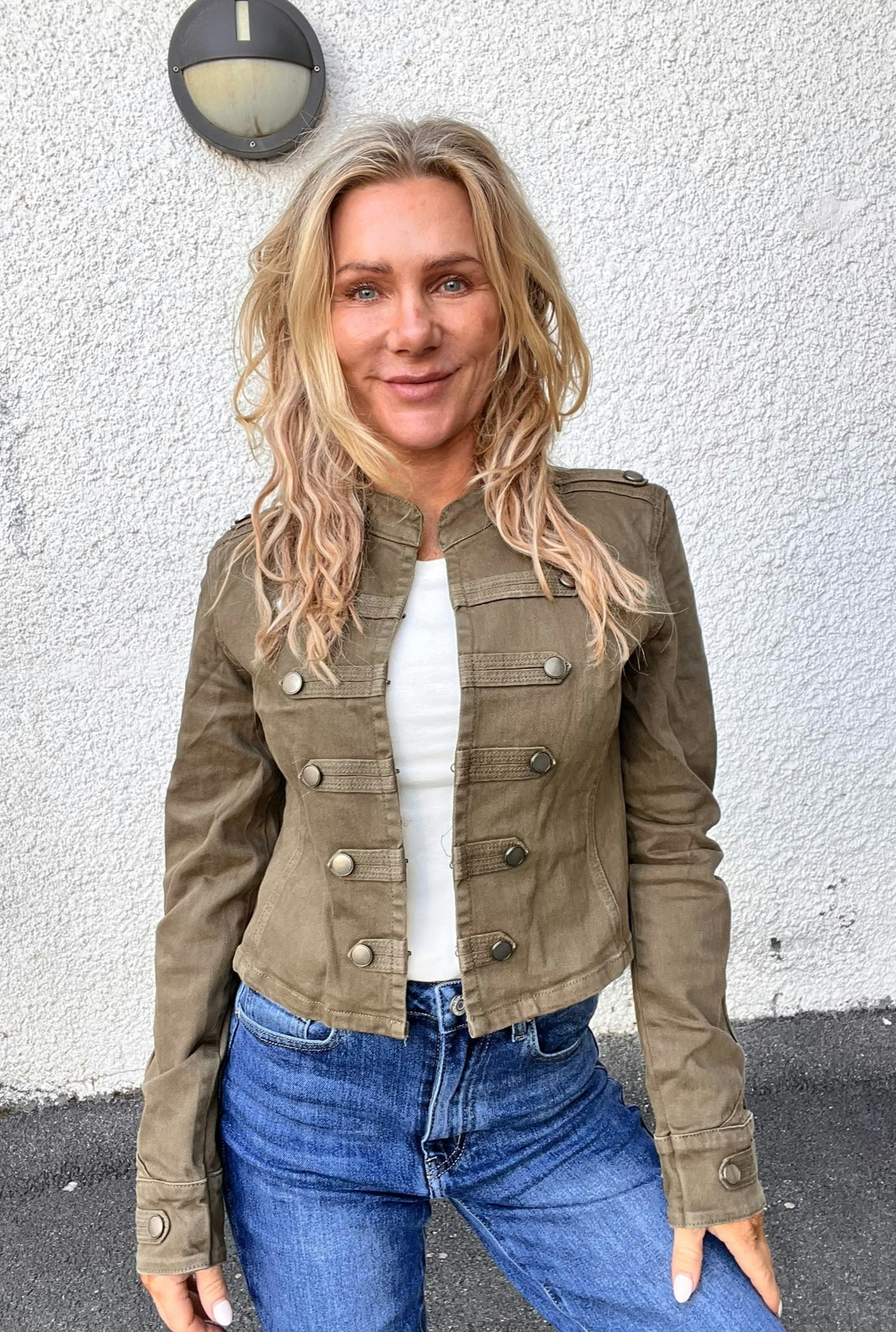 Kvinna med långt blont hår står och ler mot kameran framför en vit, grovputsad vägg med rund vägglampa ovanför. Hon bär olivgrön jacka med knappar, vit topp och blå jeans.