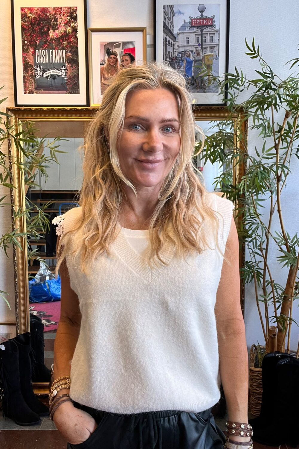 Leende kvinna med långt blont hår står framför en guldspegel och tittar mot kameran, klädd i vit stickad väst och mörka byxor. Bakom syns växter och inramade bilder med texten “CASA FANNY” och “METRO”.