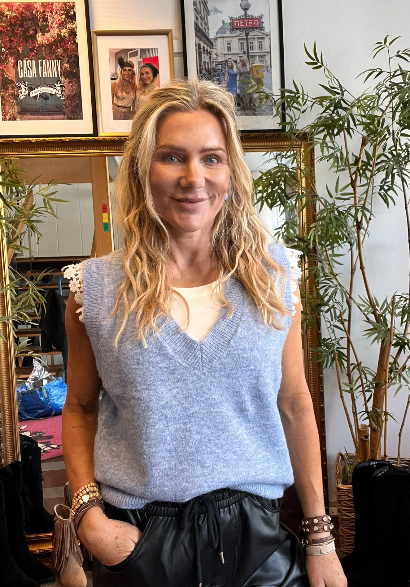 Leende kvinna med vågigt blont hår står inomhus med handen i fickan, iklädd ljusblå stickad väst och svart byxa. Bakom syns spegel, växter och inramade bilder med texten “CASA FANNY” och “METRO”.