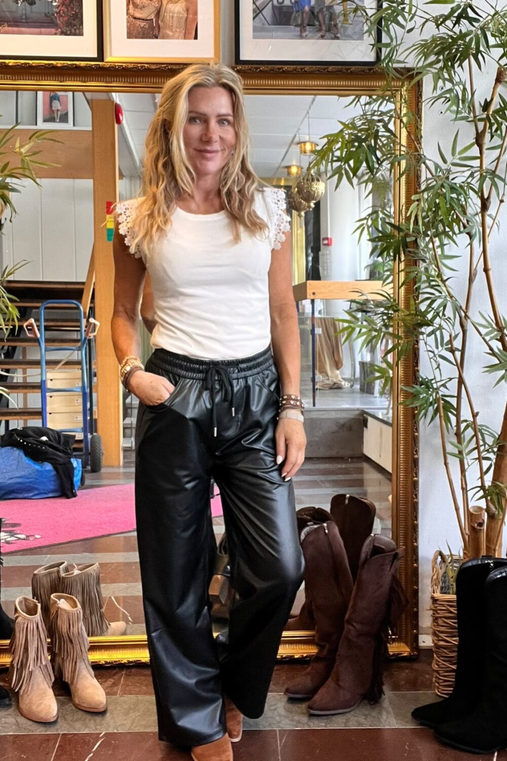 Blond kvinna poserar med ena handen i fickan framför en stor guldramsspegel, klädd i vit topp med spetsärmar och svarta läderbyxor. Runt henne syns växter, uppställda stövlar och inramade foton på väggen.
