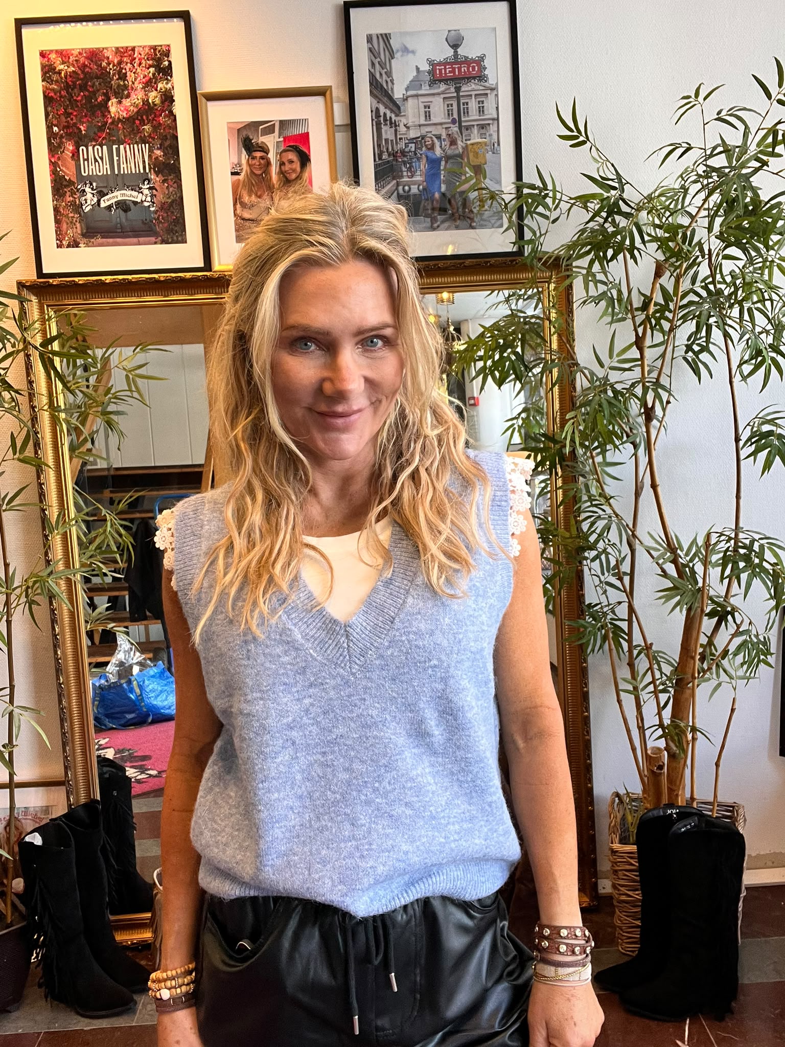 Blond kvinna står och ler mot kameran i ett rum med stora krukväxter och en guldspegel. På väggen bakom hänger inramade bilder, bland annat texten “CASA FANNY” och en metrobild med skylten “METRO”.