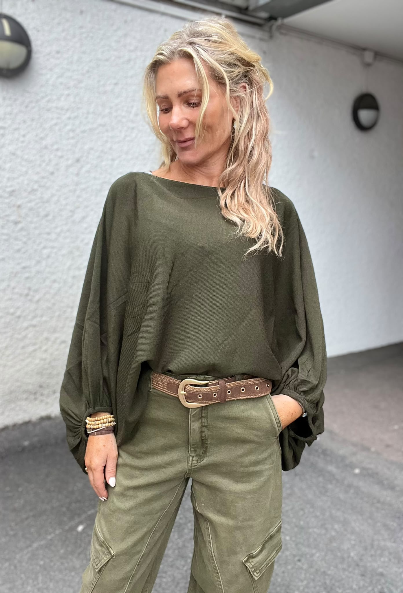 Blond kvinna står utomhus och tittar ned med ett svagt leende, ena handen i fickan. Hon bär olivgrön, lös tröja och matchande byxor med brunt bälte och armband, framför grå vägg.